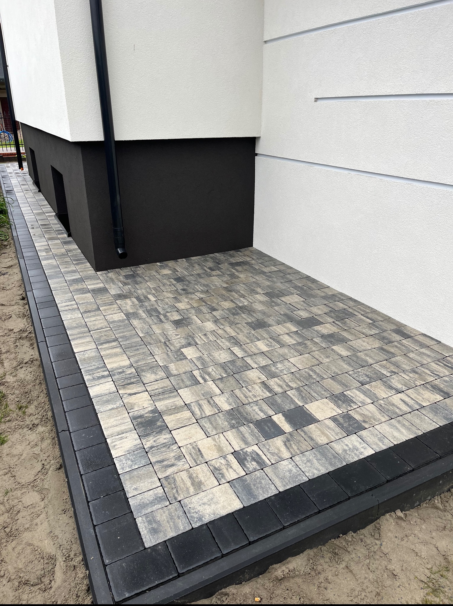 Ułożona kostka brukowa w odcieniach szarości i beżu obramowana ciemną kostką przy ścianie budynku z rynną spustową. Estetyczne wykończenie terenu przy domu.