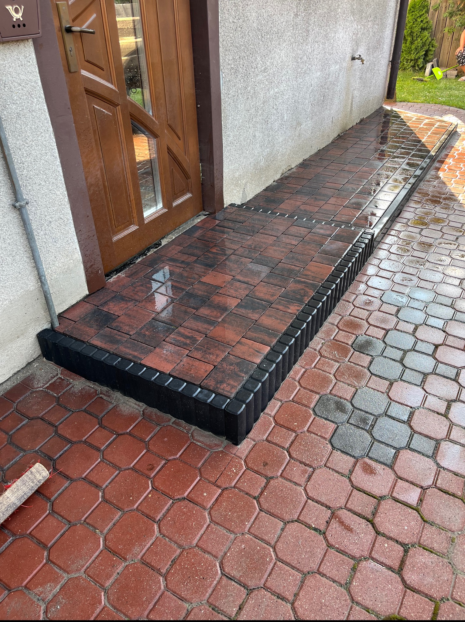 Ułożony chodnik z kostki brukowej w odcieniach czerwieni i czerni, prowadzący do drzwi wejściowych. Widoczne obrzeża i elementy drenażowe.