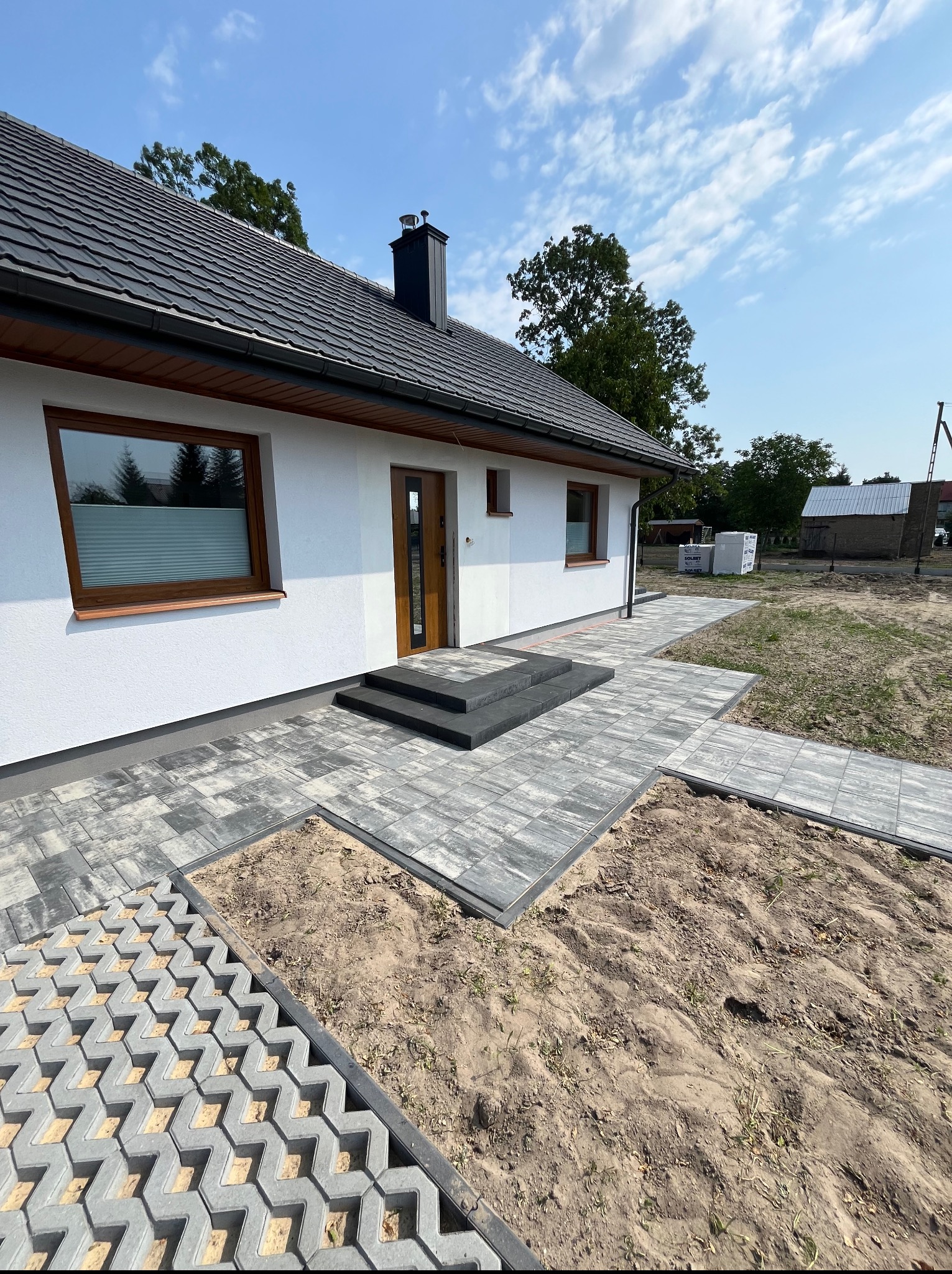 Nowoczesny podjazd z kostki brukowej w odcieniach szarości prowadzi do domu z brązowymi oknami. Część terenu wyłożona ażurową kostką. Słoneczny dzień.