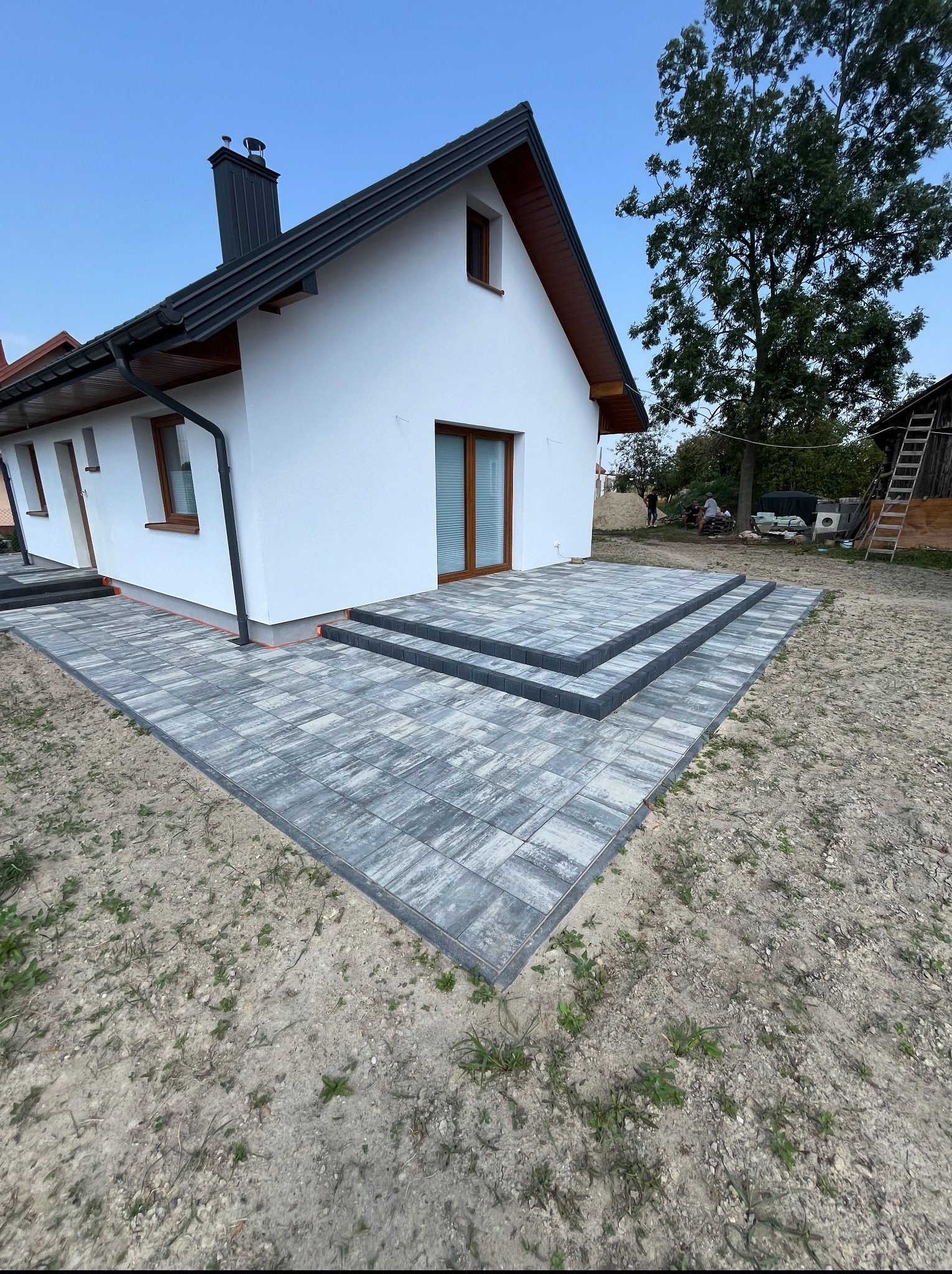 Nowoczesny taras z szarej kostki brukowej przed białym domem z ciemnymi oknami i dachem. Widoczne schody z kostki i fragment ogrodu.