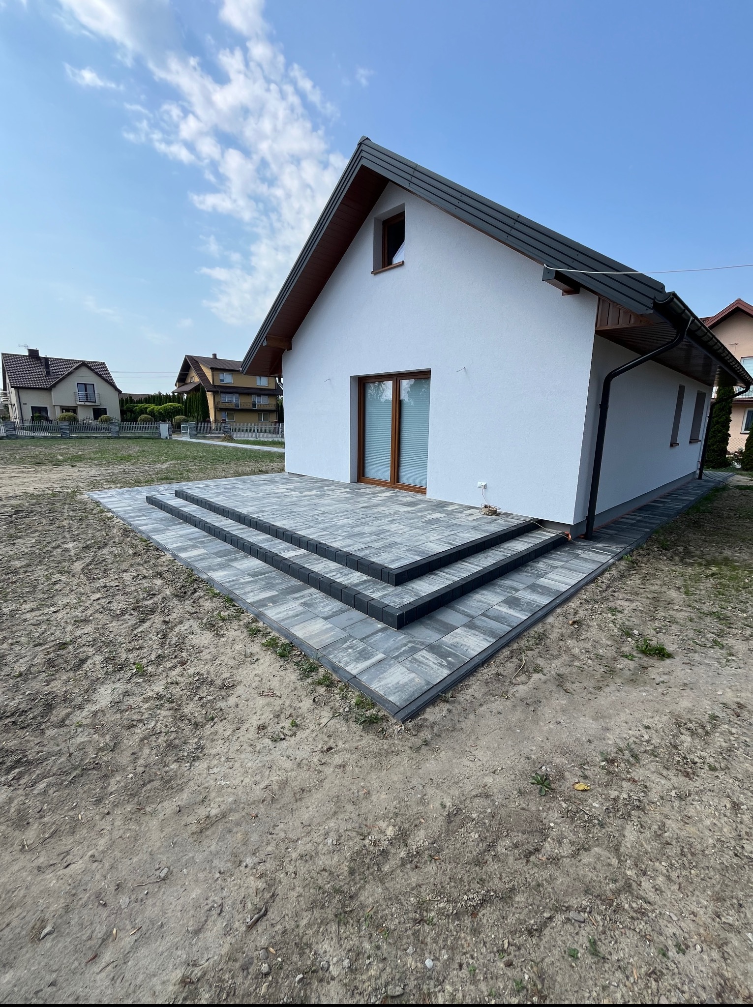 Nowoczesny taras z kostki brukowej w odcieniach szarości, dopasowany do elewacji domu jednorodzinnego. Widok na starannie ułożone stopnie i obrzeża, trawnik w tle.