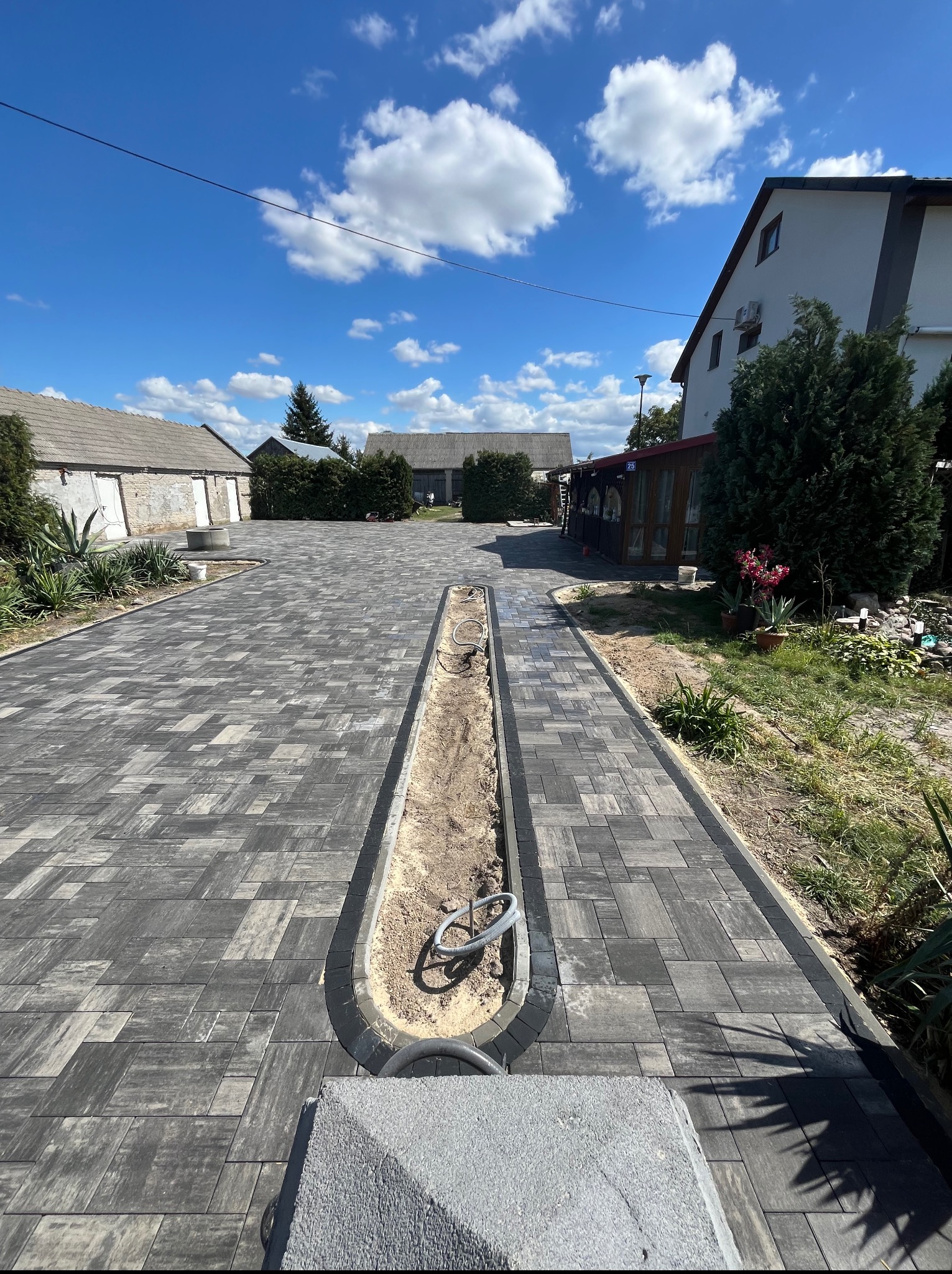 Ułożona szara kostka brukowa na podjeździe z elementami drenażu liniowego, widok na budynki w tle, słoneczny dzień.