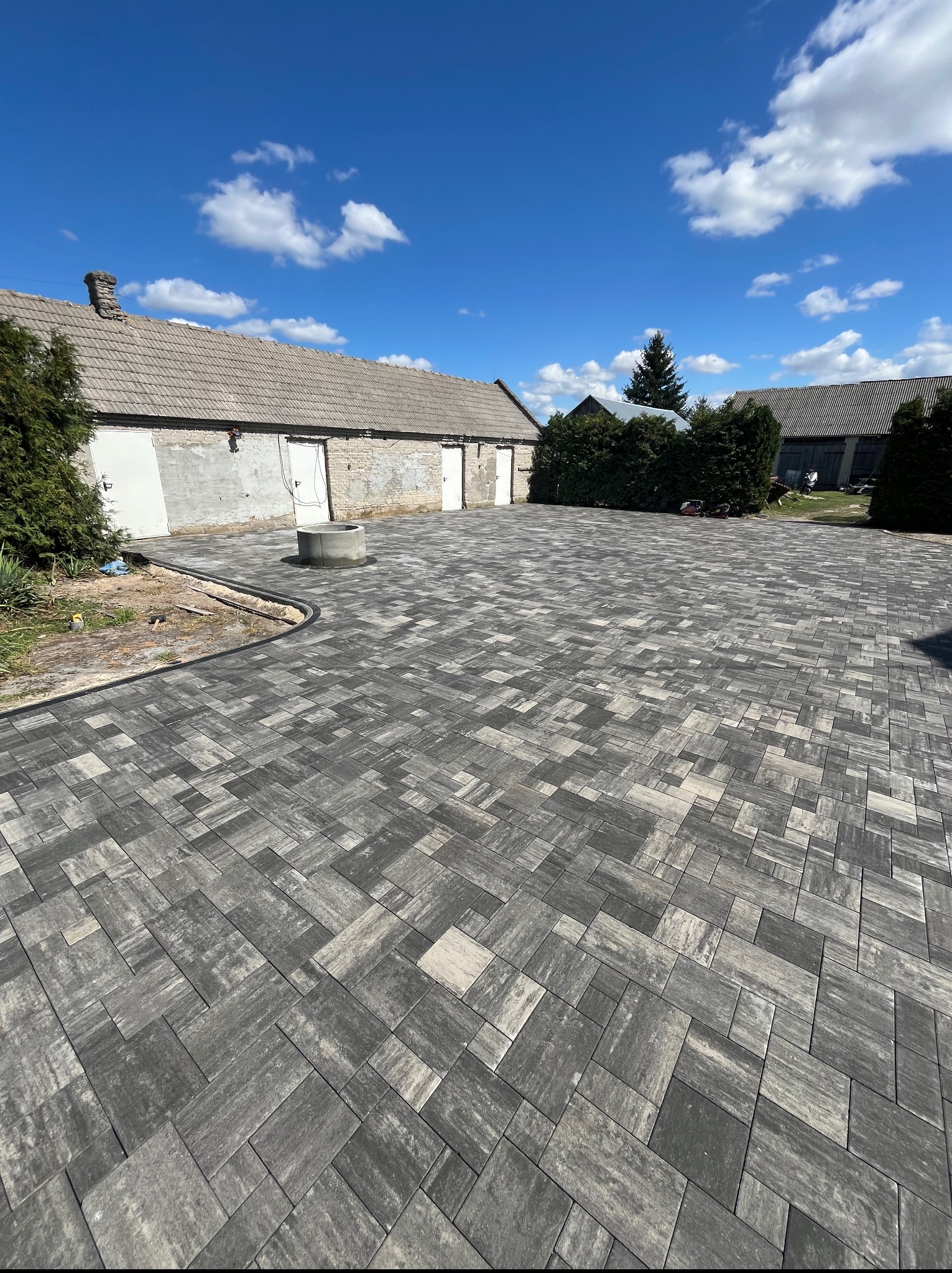 Ułożona szara kostka brukowa na podwórzu z widocznym budynkiem gospodarczym i studnią. Nowa nawierzchnia z obrzeżami, słoneczny dzień.