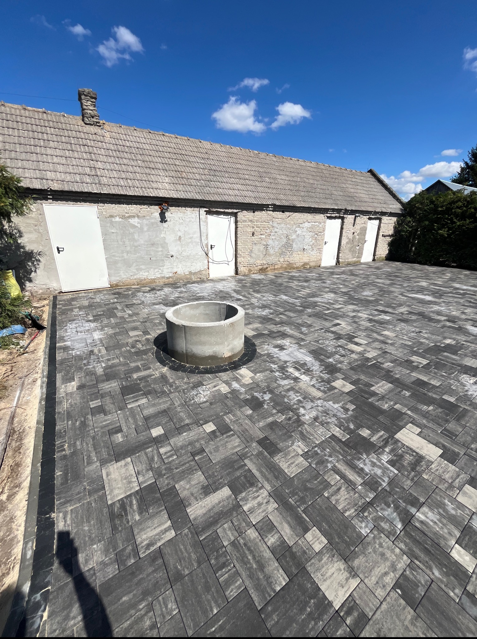 Ułożona kostka brukowa w odcieniach szarości z betonową studzienką na dziedzińcu, na tle budynku z białymi drzwiami i błękitnego nieba.