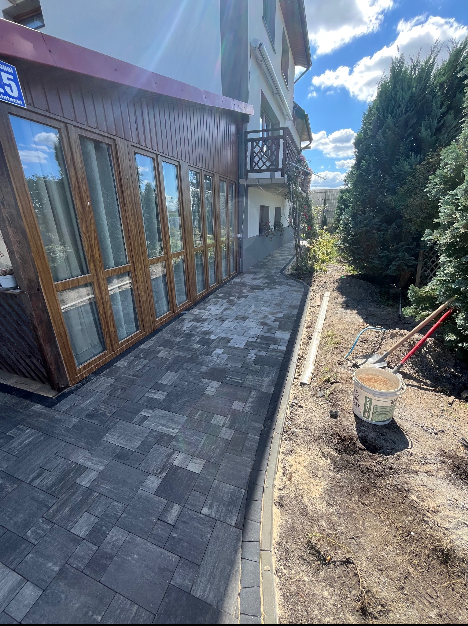 Ścieżka z szarej kostki brukowej prowadząca wzdłuż domu z drewnianą werandą, obok narzędzia i niezagospodarowany teren. Słoneczny dzień.