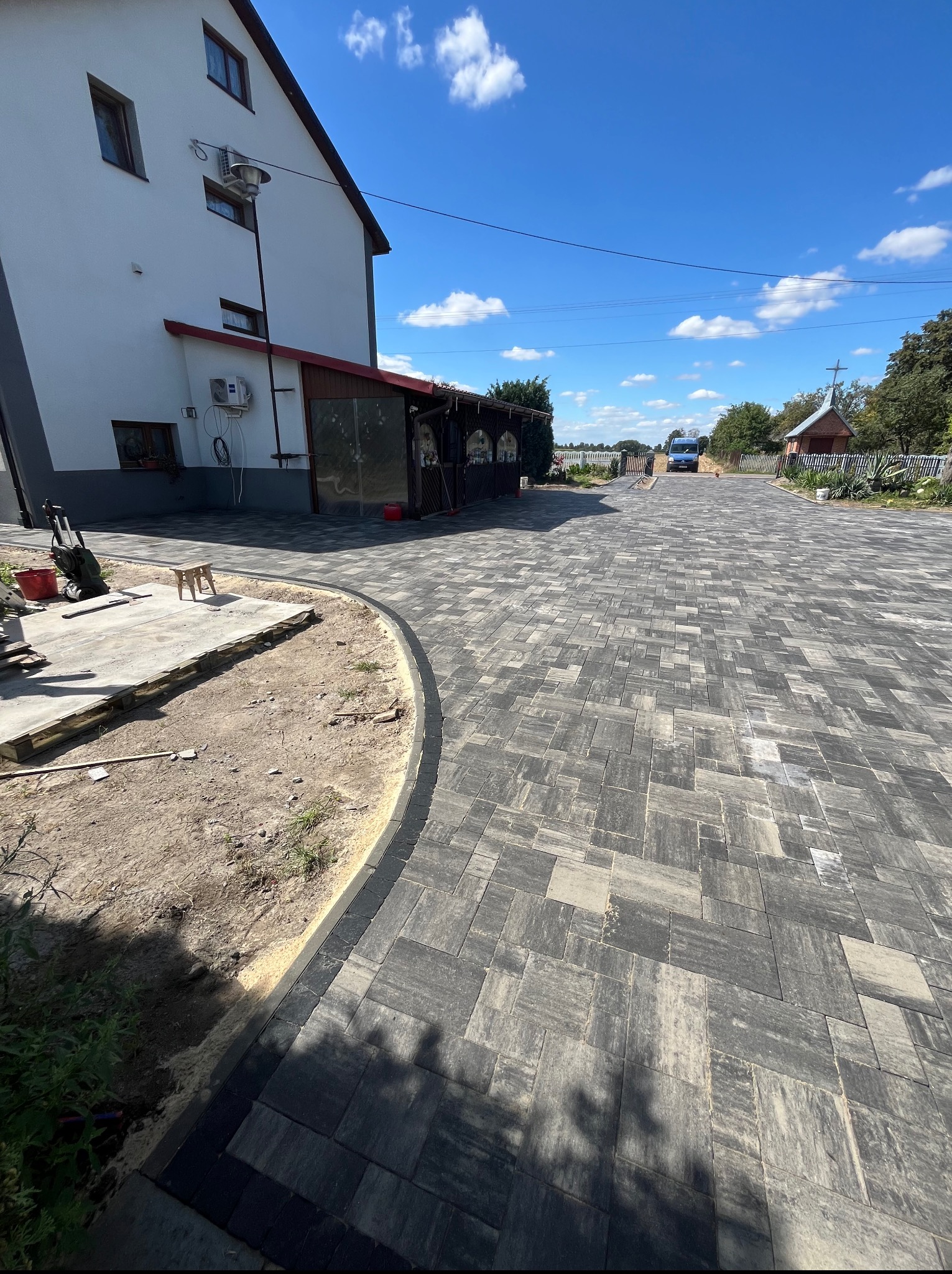 Szeroki podjazd z kostki brukowej w odcieniach szarości, zakończony obrzeżem. Częściowo ułożona kostka przy budynku. Błękitne niebo w tle.