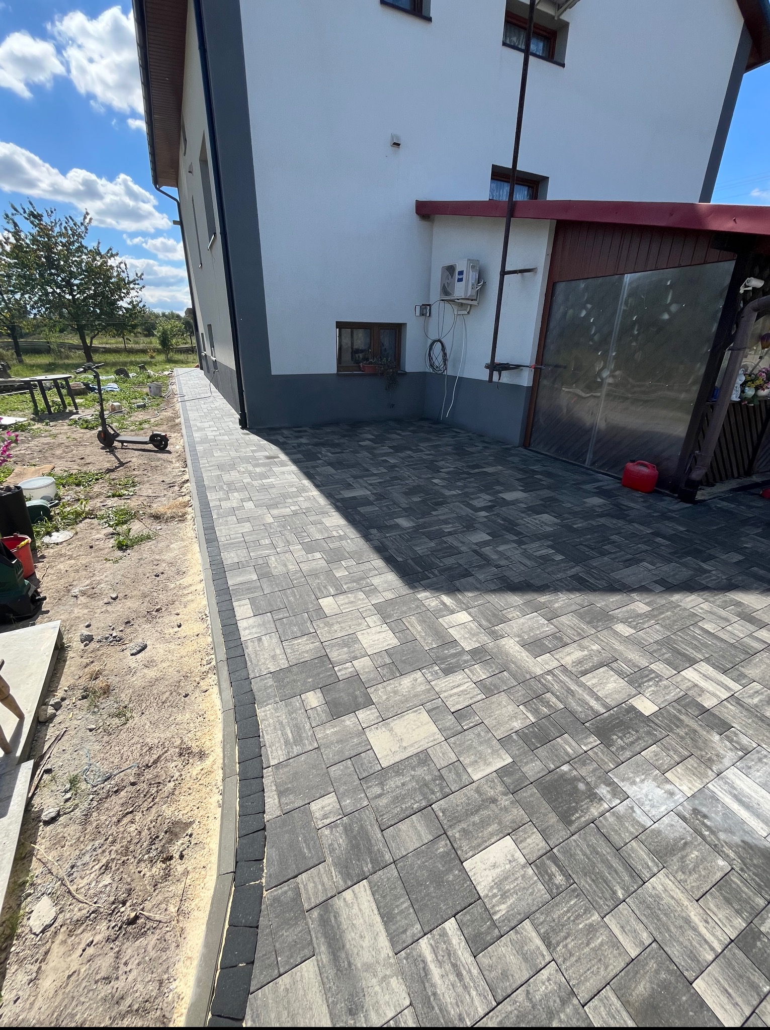 Ułożona kostka brukowa w szaro-białej kolorystyce przy domu z widocznym obrzeżem i przygotowanym terenem pod dalsze prace.