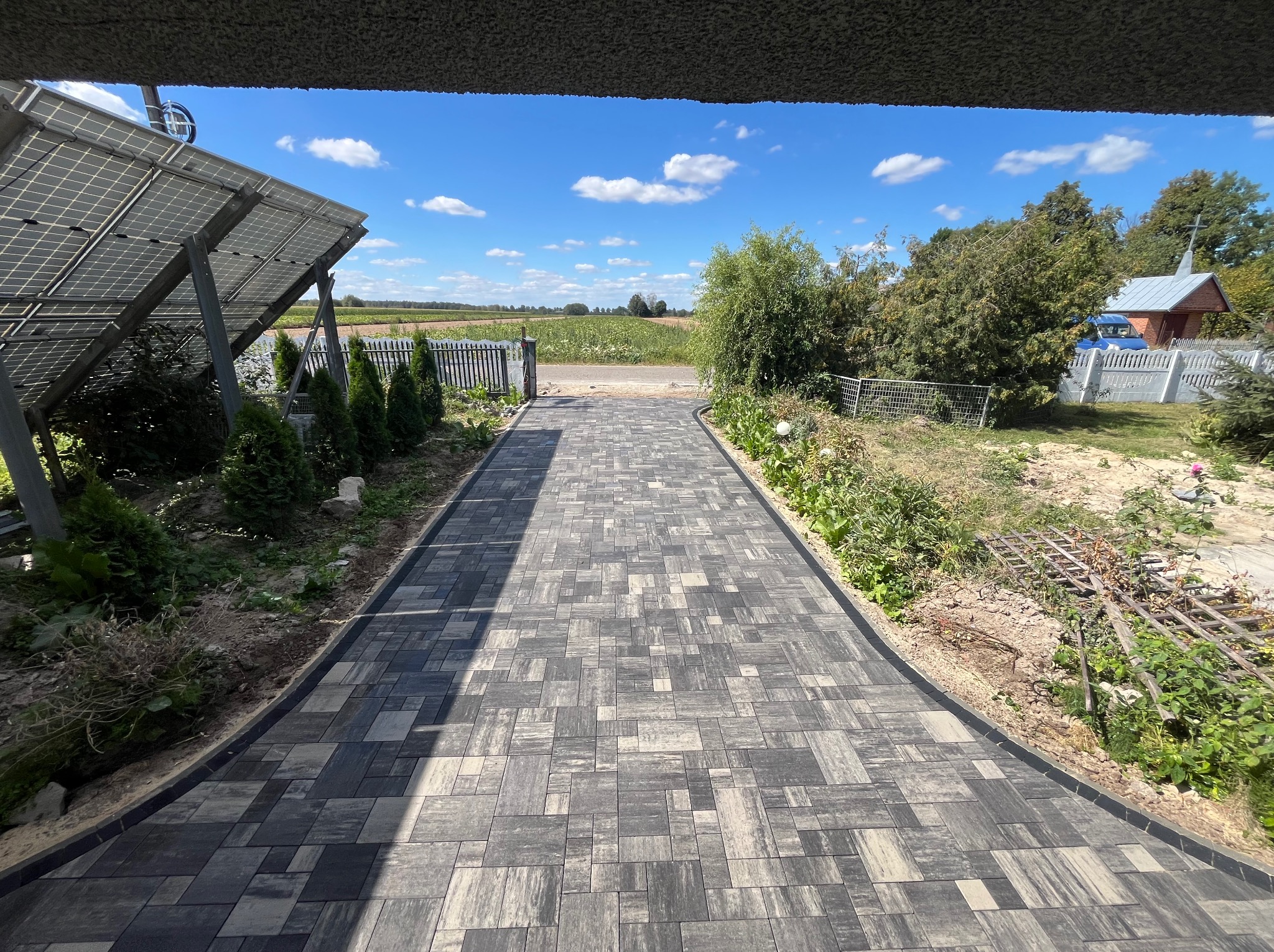 Szeroki, wybrukowany podjazd z kostki w odcieniach szarości prowadzi do bramy w tle, obok paneli słonecznych po lewej i zieleni po prawej, pod zadaszeniem.