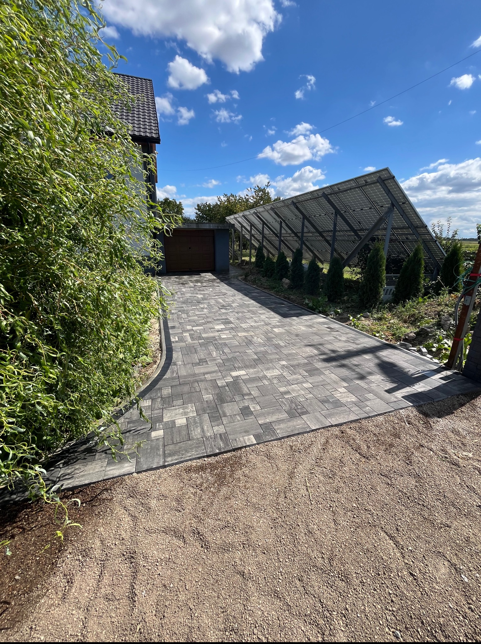 Ułożona, szara kostka brukowa prowadząca do garażu pod panele słoneczne. Krawężnik oddziela kostkę od żwirowego podłoża. W tle błękitne niebo z chmurami.