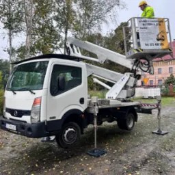 Podnośnik koszowy na mokrym terenie, z operatorem w żółtej kamizelce. Widoczny budynek w tle i drzewa. Tablica informacyjna z ofertą usług wysokościowych.