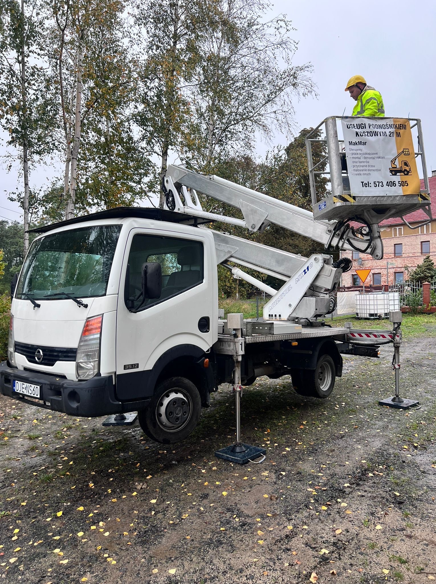 Podnośnik koszowy na mokrym terenie, z operatorem w żółtej kamizelce. Widoczny budynek w tle i drzewa. Tablica informacyjna z ofertą usług wysokościowych.
