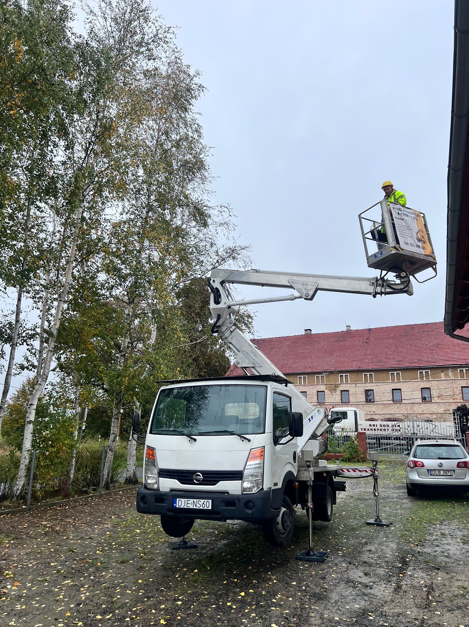 Biały podnośnik koszowy na nierównym terenie, z operatorem w żółtym kasku w koszu. W tle budynek z czerwonym dachem i drzewa z jesiennymi liśćmi.
