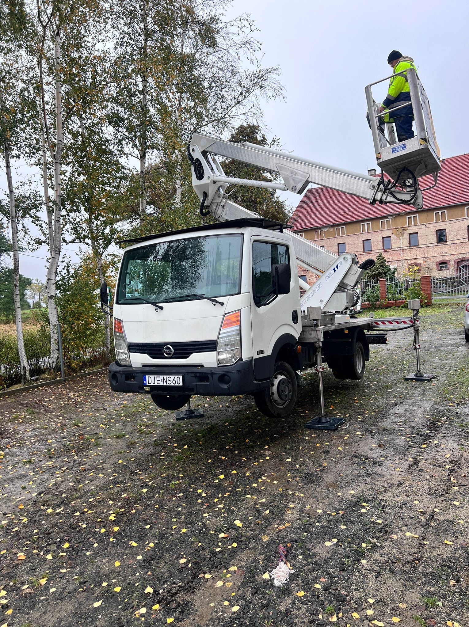 Biały podnośnik koszowy Multitel MX210 na mokrym gruncie z opadłymi liśćmi, pracownik w koszu na tle ceglanego budynku i drzew.