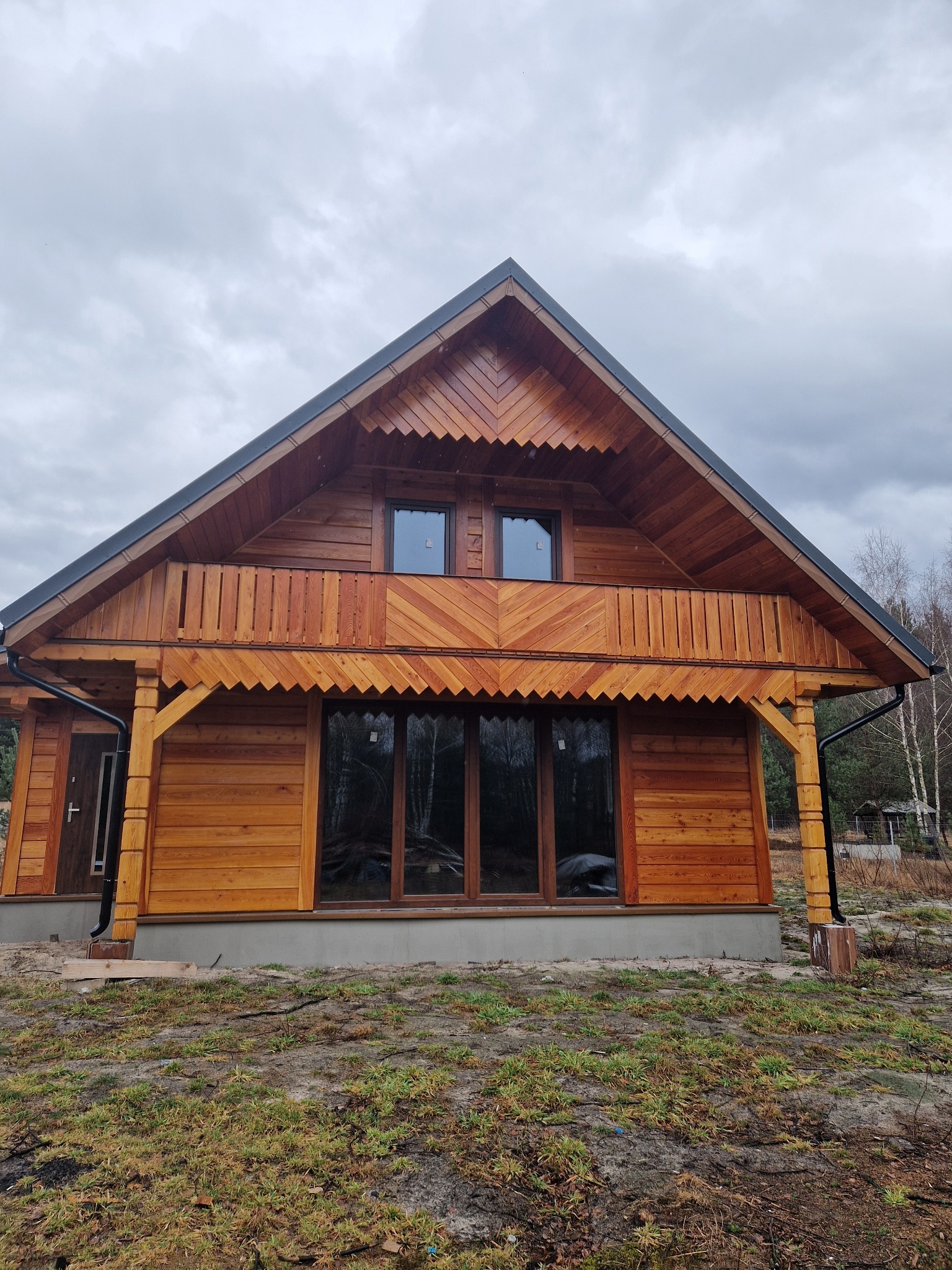 Drewniany dom z balkonem w Krasnobrodzie, z charakterystycznymi zdobieniami pod okapem i na balustradzie, na tle pochmurnego nieba i lasu.