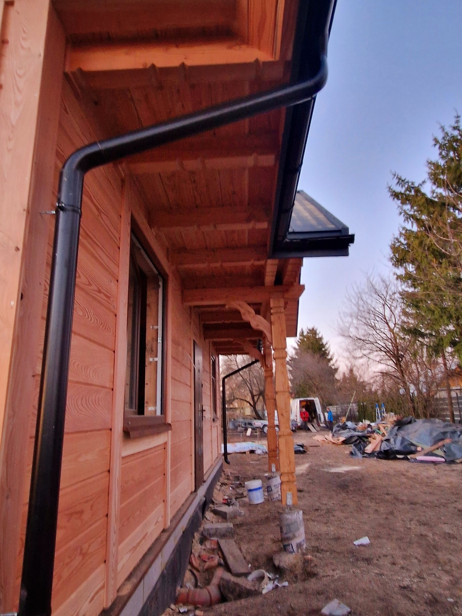 Wykończona elewacja domu z drewna, z czarną rynną i stolarką okienną. Widoczny fragment werandy i otoczenie w trakcie prac wykończeniowych.