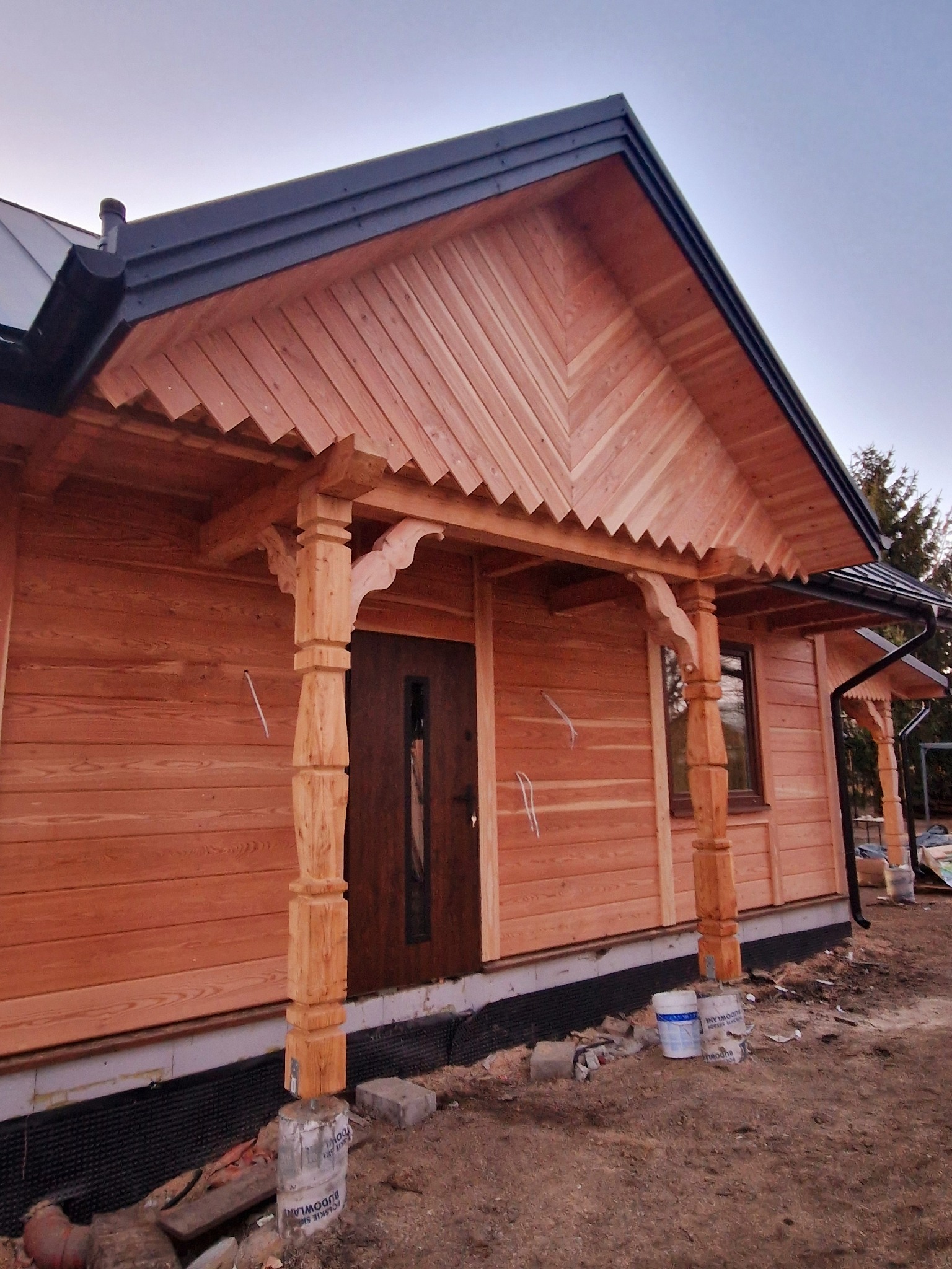 Surowy dom z bali z drewnianym gankiem i kolumnami, widoczne detale wykończenia dachu i elewacji. Na pierwszym planie materiały budowlane.