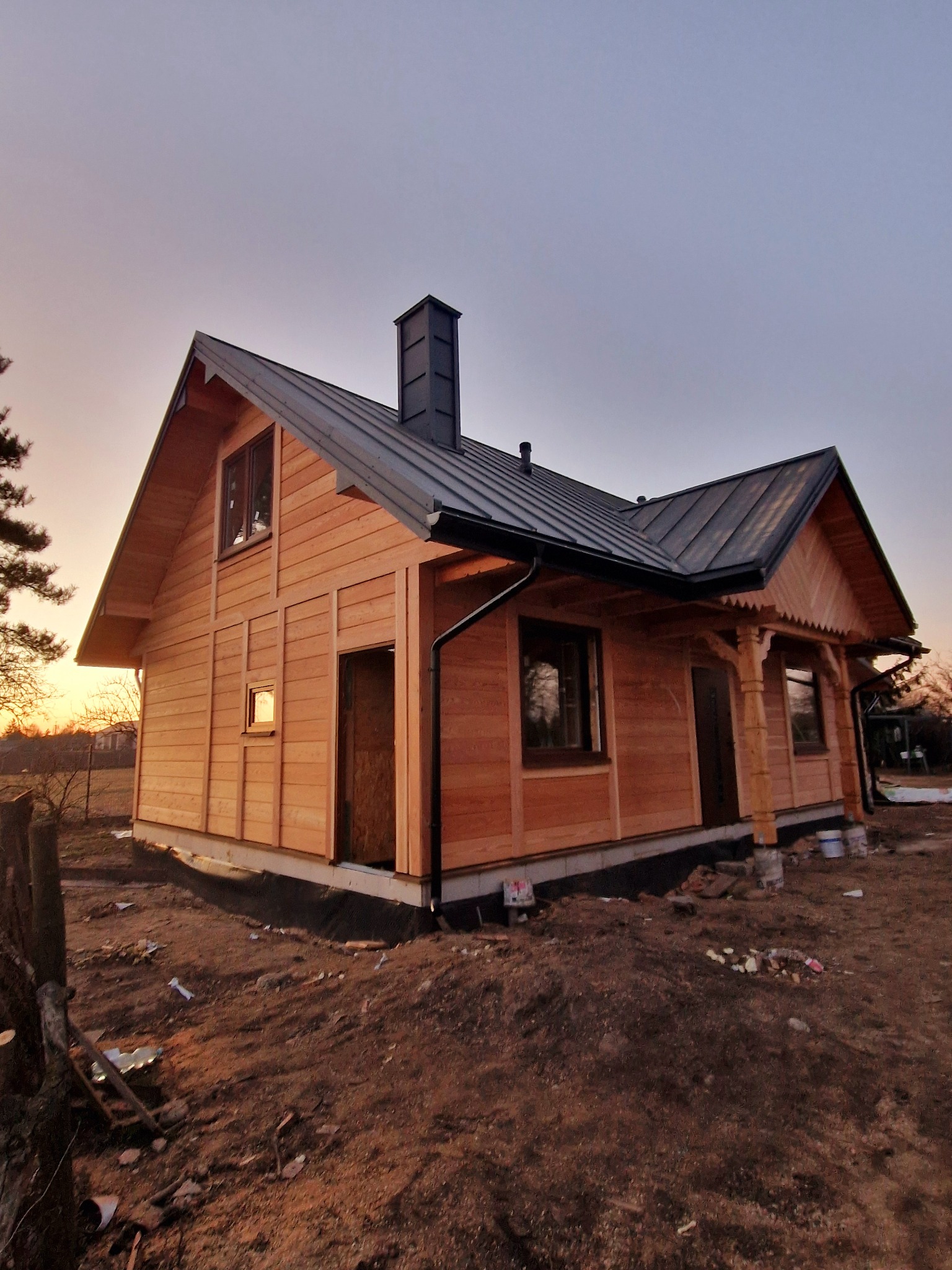 Wykończony drewniany dom z czarnym dachem i kominem, widziany pod kątem o zmierzchu. Widoczne detale drewnianej elewacji i elementy wykończeniowe.