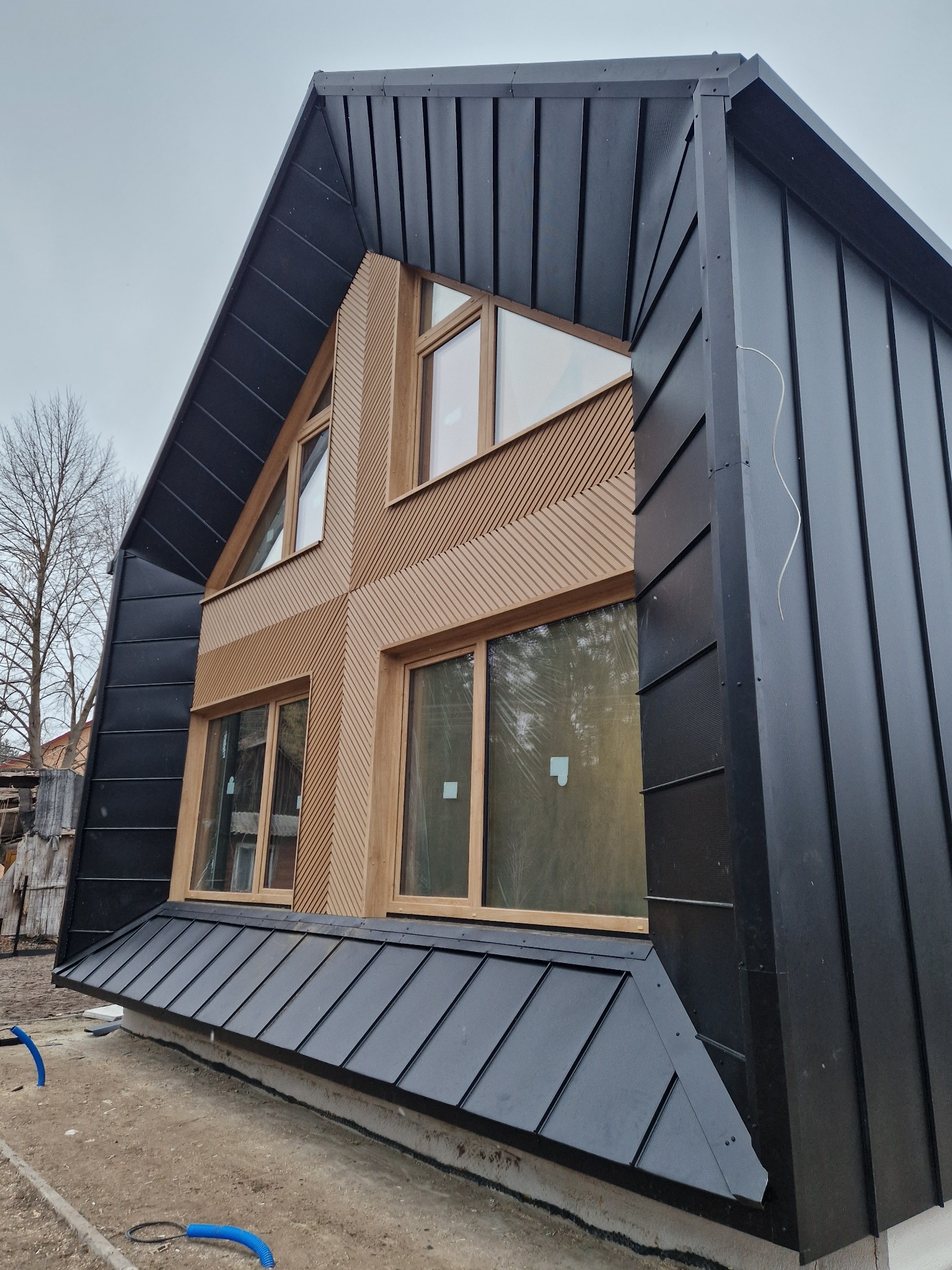 Nowoczesny dom szkieletowy z czarnym metalowym dachem i elewacją z drewnianymi panelami. Charakterystyczne okna z drewnianymi ramami. Surowy stan wykończenia.