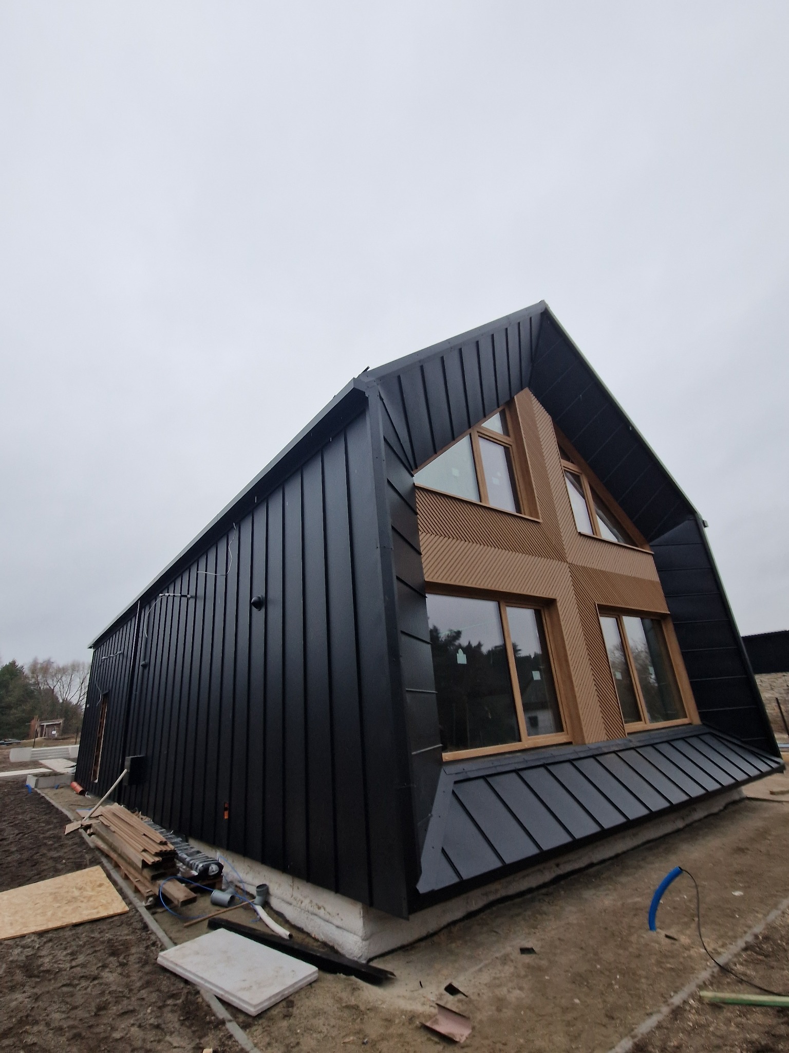 Czarny dom szkieletowy o unikalnej bryle z drewnianymi akcentami i oknami w kształcie krzyża, w trakcie budowy, widok z dołu na tle pochmurnego nieba.