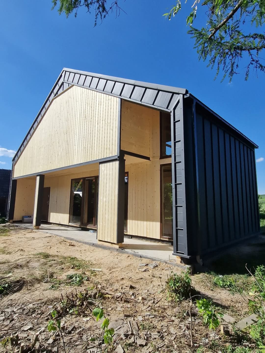 Surowy, nowoczesny dom z drewna i blachy na etapie budowy, z widoczną konstrukcją i dużymi oknami, w słoneczny dzień. Detal: widoczne łączenia elementów.