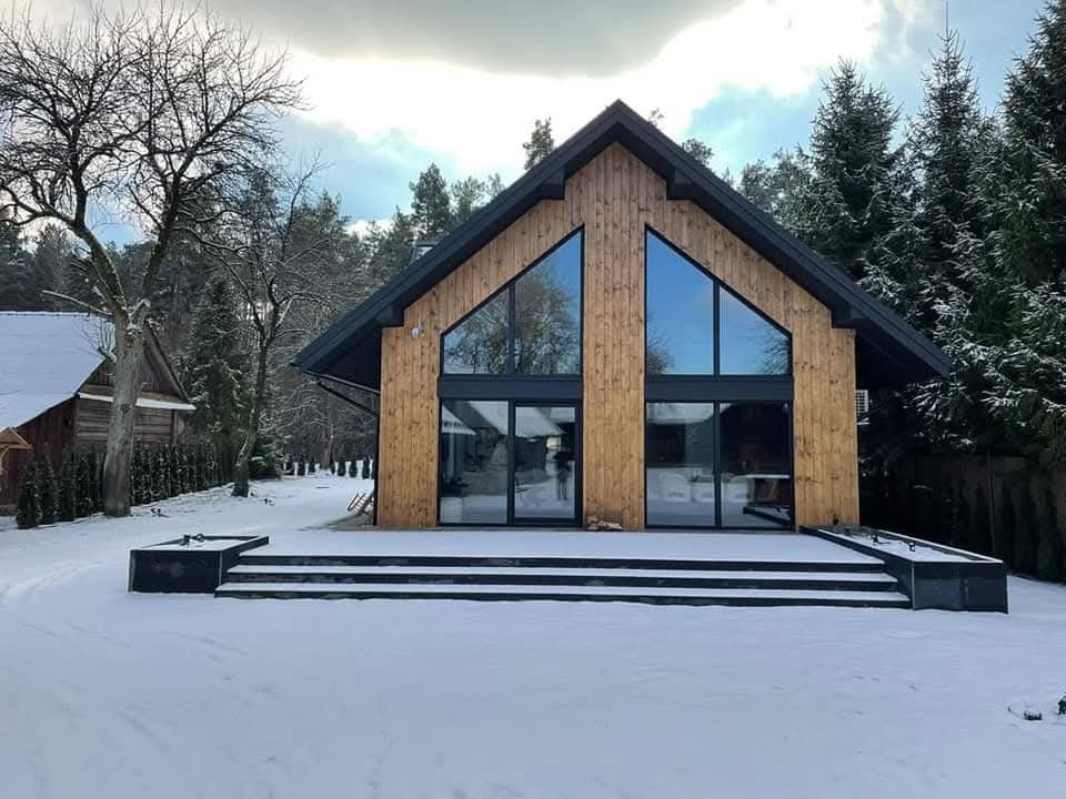 Nowoczesny dom z drewna i szkła, z dużymi oknami trójkątnymi, otoczony śniegiem i drzewami. Architektura w stylu skandynawskim, z czarnymi akcentami i drewnianą elewacją.