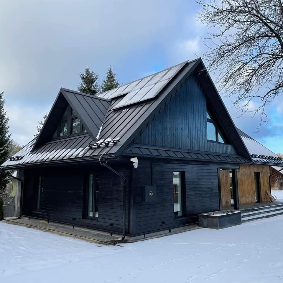 Nowoczesny, czarny dom z drewna z panelami słonecznymi na dachu, otoczony śniegiem. Widoczne detale wykończenia i elementy architektury.