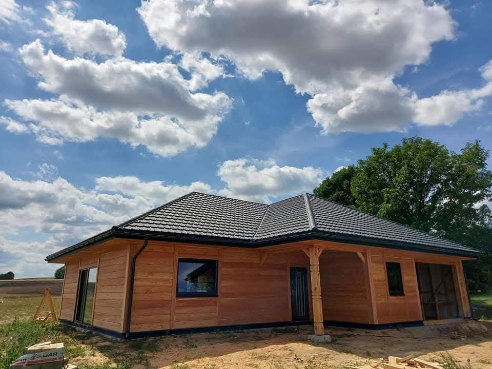 Parterowy budynek z drewnianą elewacją i ciemnym dachem, z widocznymi słupami podtrzymującymi zadaszenie werandy, na tle błękitnego nieba z chmurami.