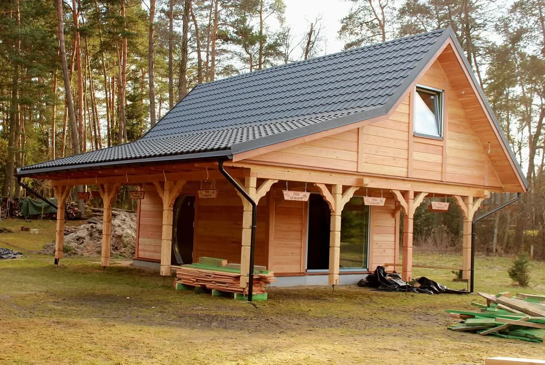 Jasny, parterowy dom szkieletowy z ciemną, metalową dachówką i zadaszonym tarasem. Na pierwszym planie stos desek i resztki materiałów budowlanych.