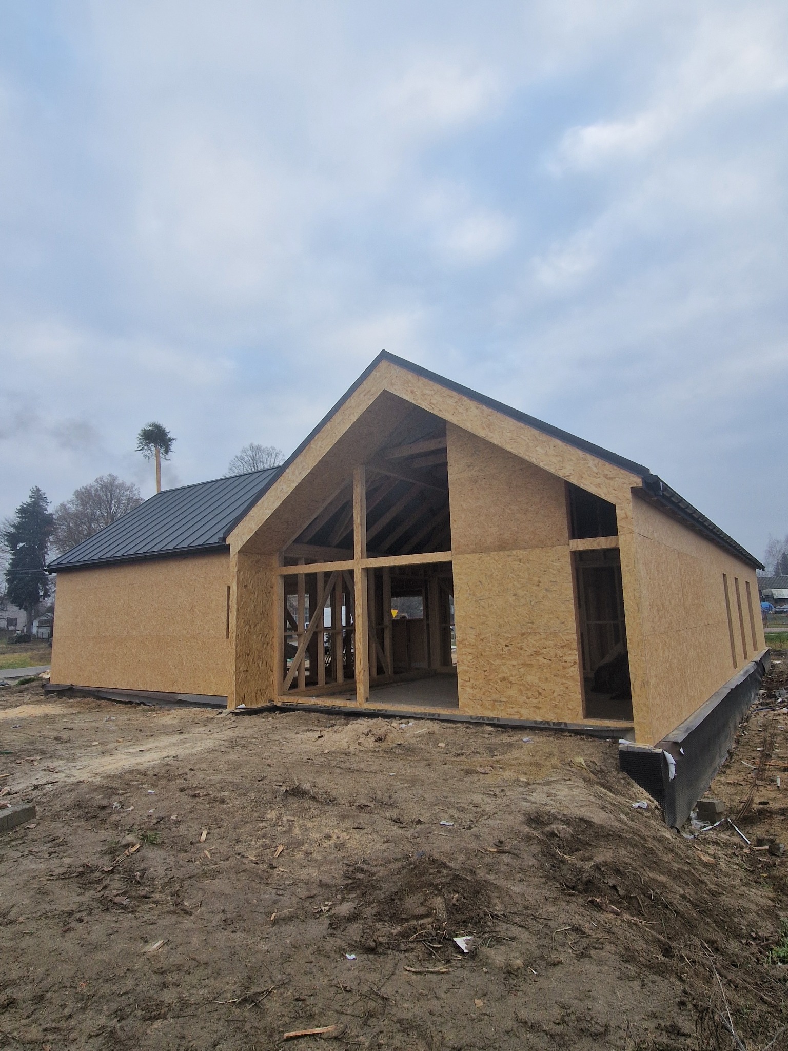 Surowy stan domu z drewnianą konstrukcją szkieletową i czarnym dachem, widok z zewnątrz na tle pochmurnego nieba. Na pierwszym planie ziemia.