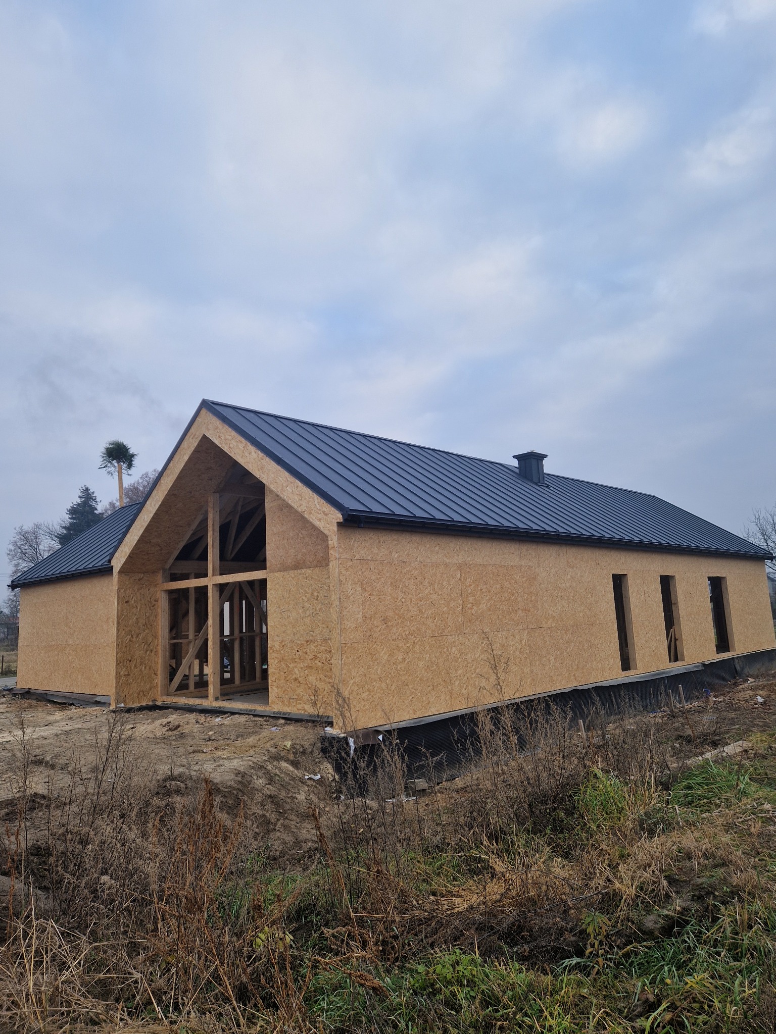 Surowy stan domu z drewnianą konstrukcją i czarnym dachem, widoczne otwory okienne. Otoczenie to ziemia i trawa, niebo pochmurne. Widok z zewnątrz.