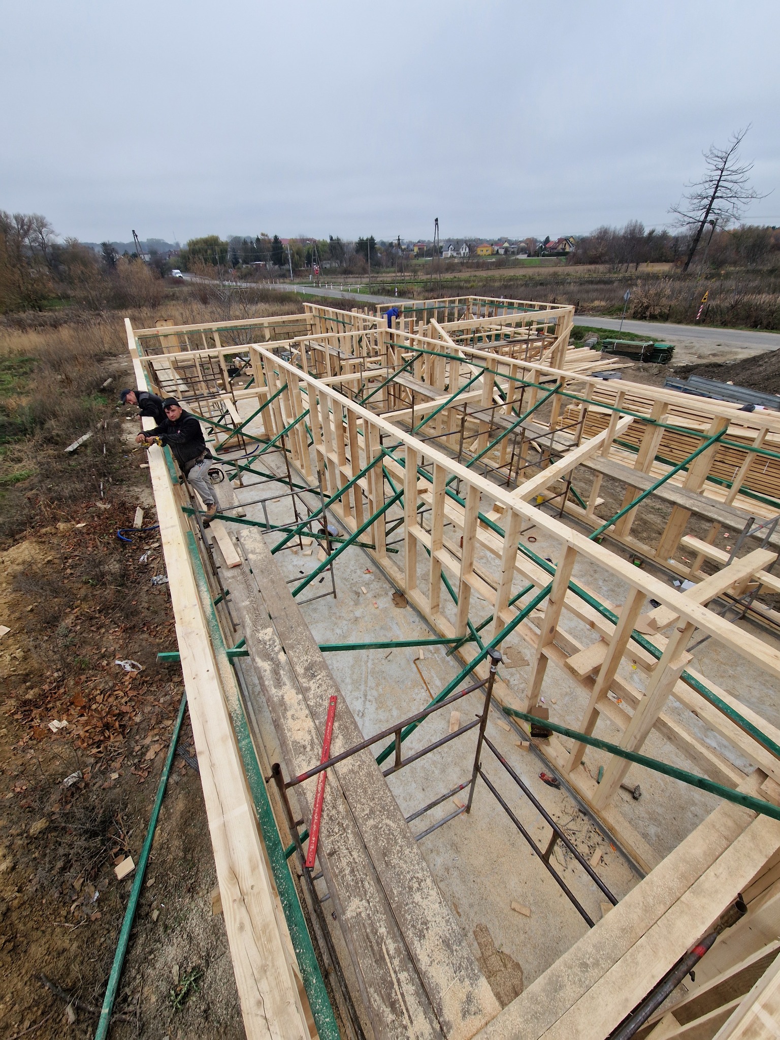 Konstrukcja domu szkieletowego w trakcie budowy z pracownikami na górnej belce. Widok z góry, zielone podpory, pochmurne niebo w tle.