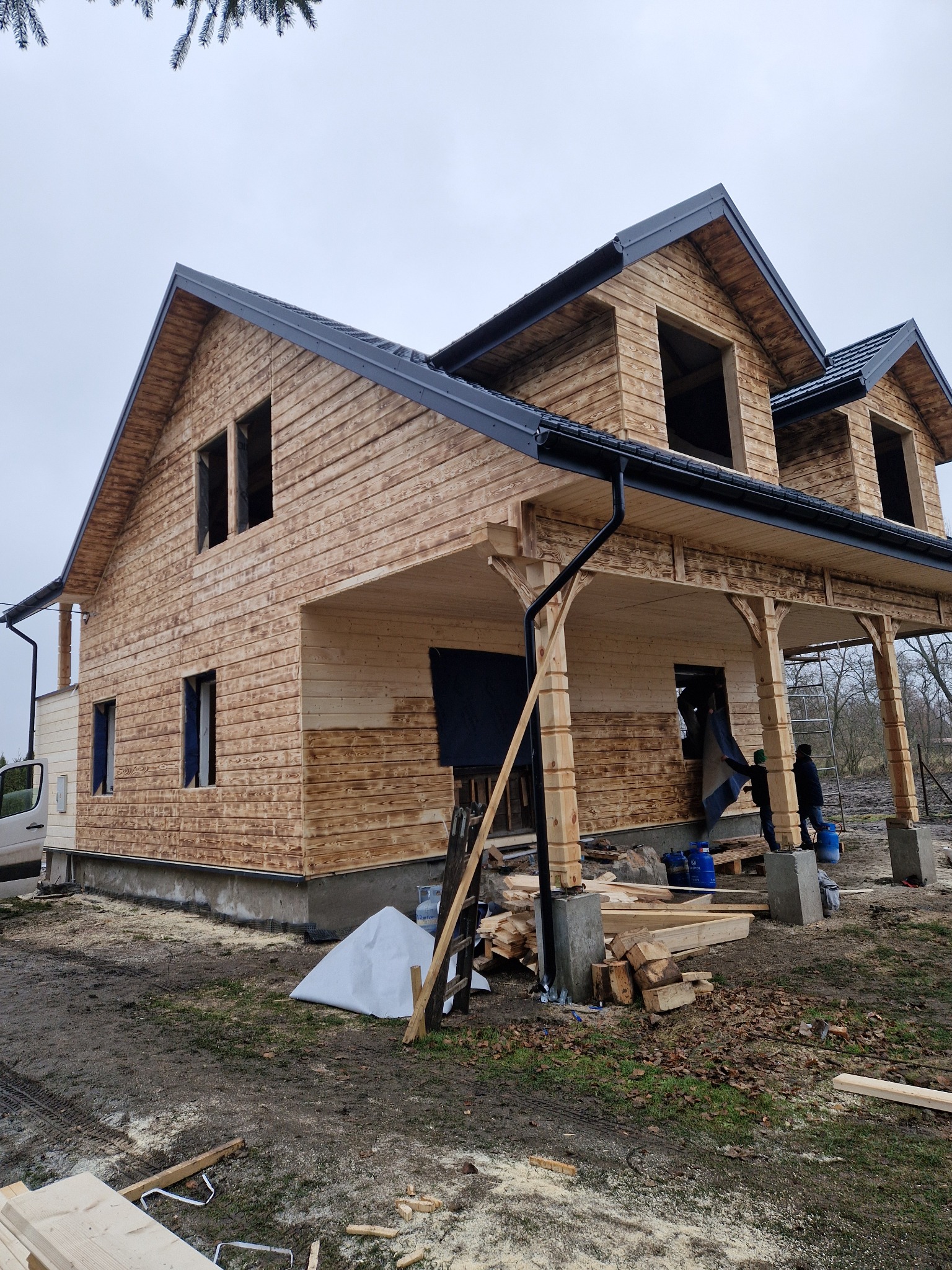 Dom szkieletowy w trakcie budowy: widoczne drewniane ściany, czarny dach, fundamenty i pracownicy montujący elementy. Częściowo wykończona elewacja.