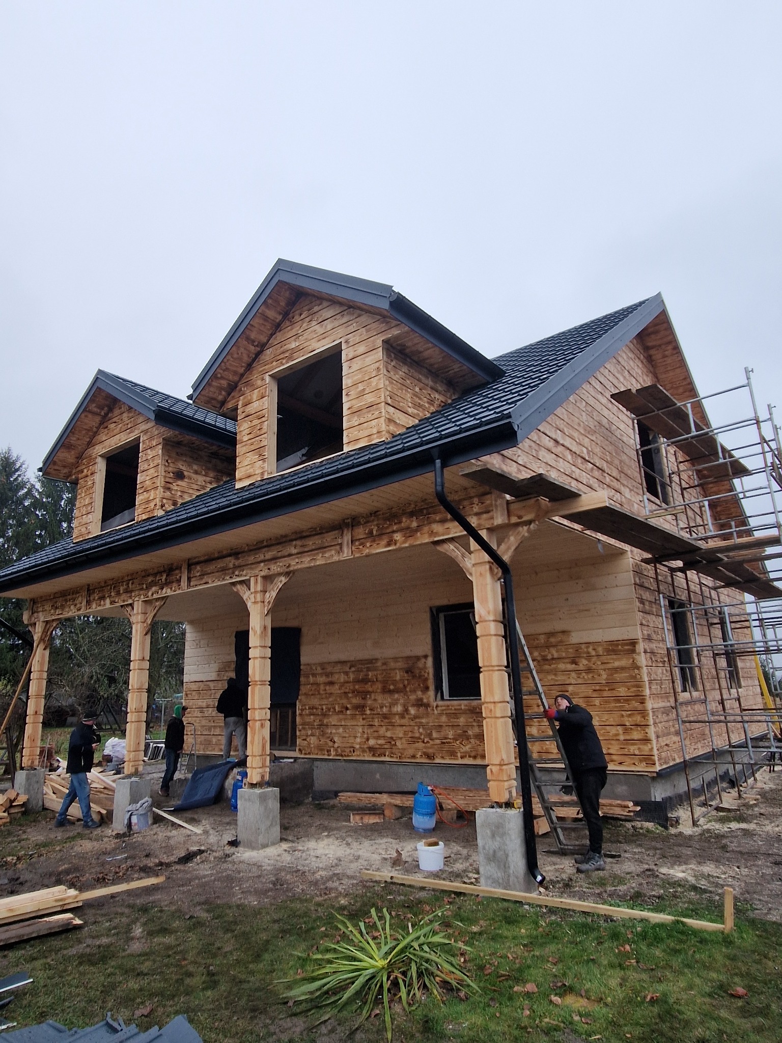 Budowa domu szkieletowego z drewnianą elewacją i czarnym dachem. Widoczne rusztowania, pracownicy i elementy konstrukcyjne. Etap impregnacji drewna.