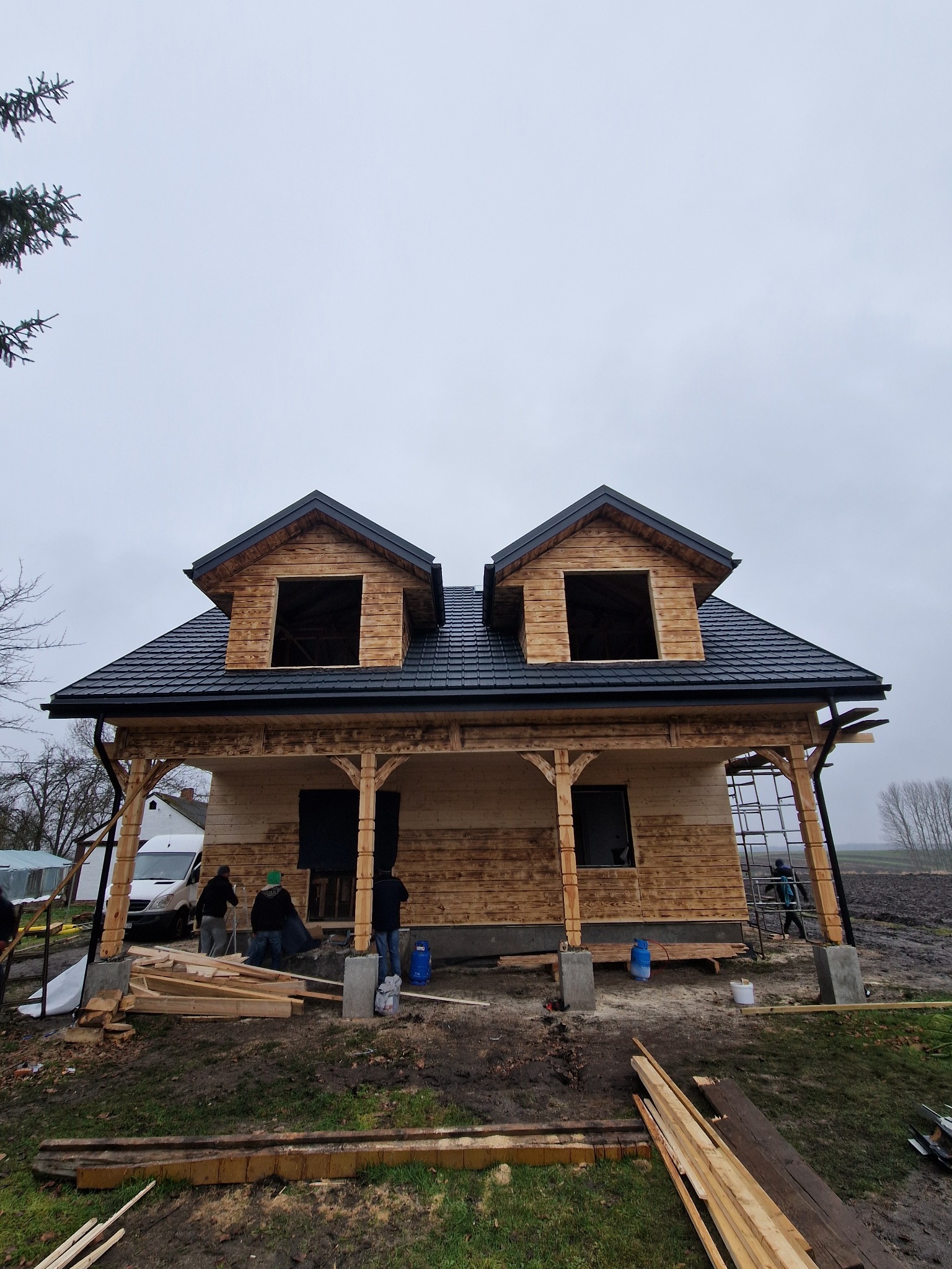 Wznoszony dom szkieletowy z ciemną dachówką, widoczne drewniane elementy konstrukcyjne i ekipa budowlana. Surowy stan, otoczenie placu budowy.