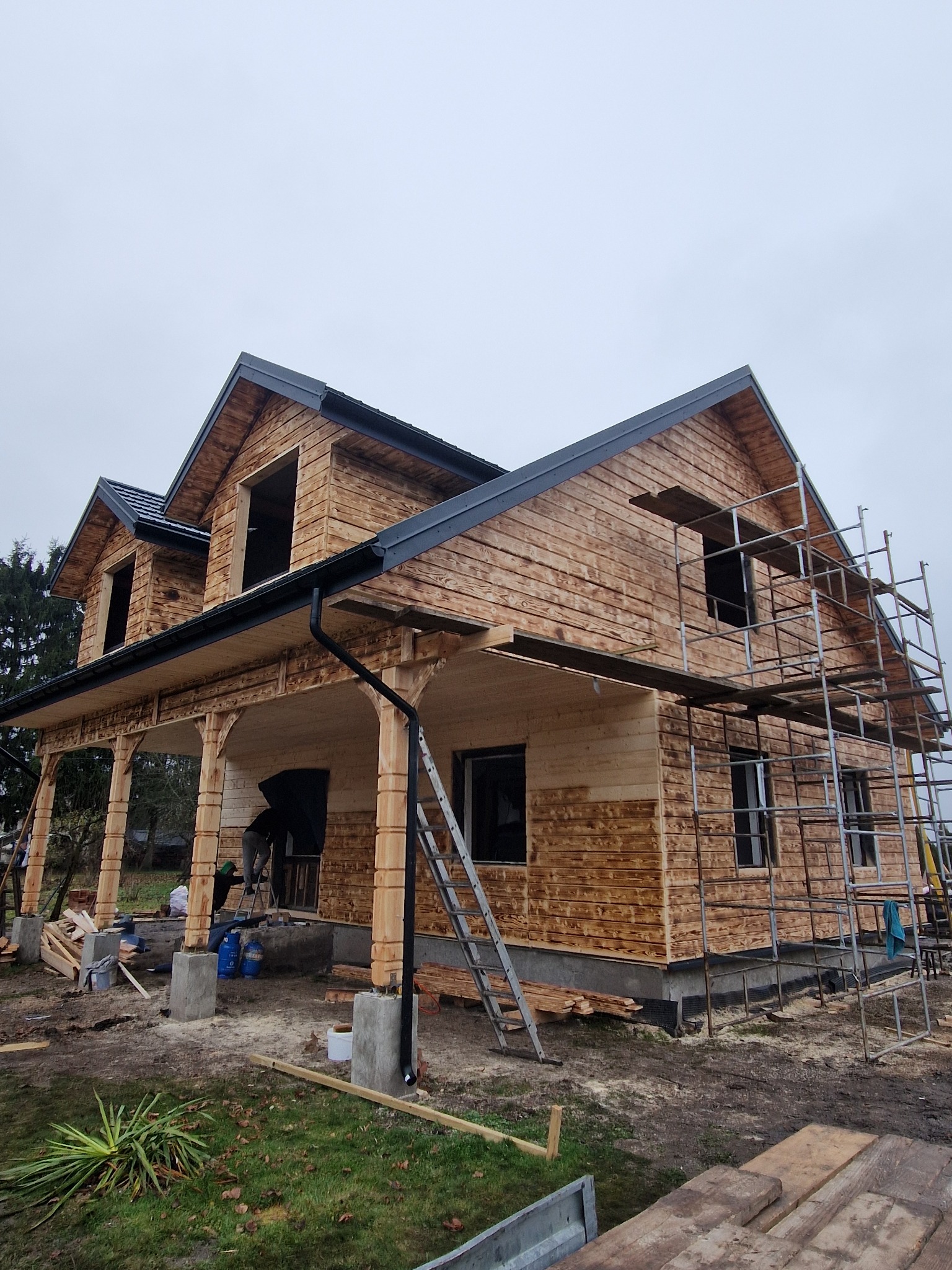 Surowy, dwukondygnacyjny dom z bali w trakcie budowy. Widoczne rusztowania, niezabudowane okna i elementy konstrukcyjne werandy. Drewno impregnowane, szara dachówka.