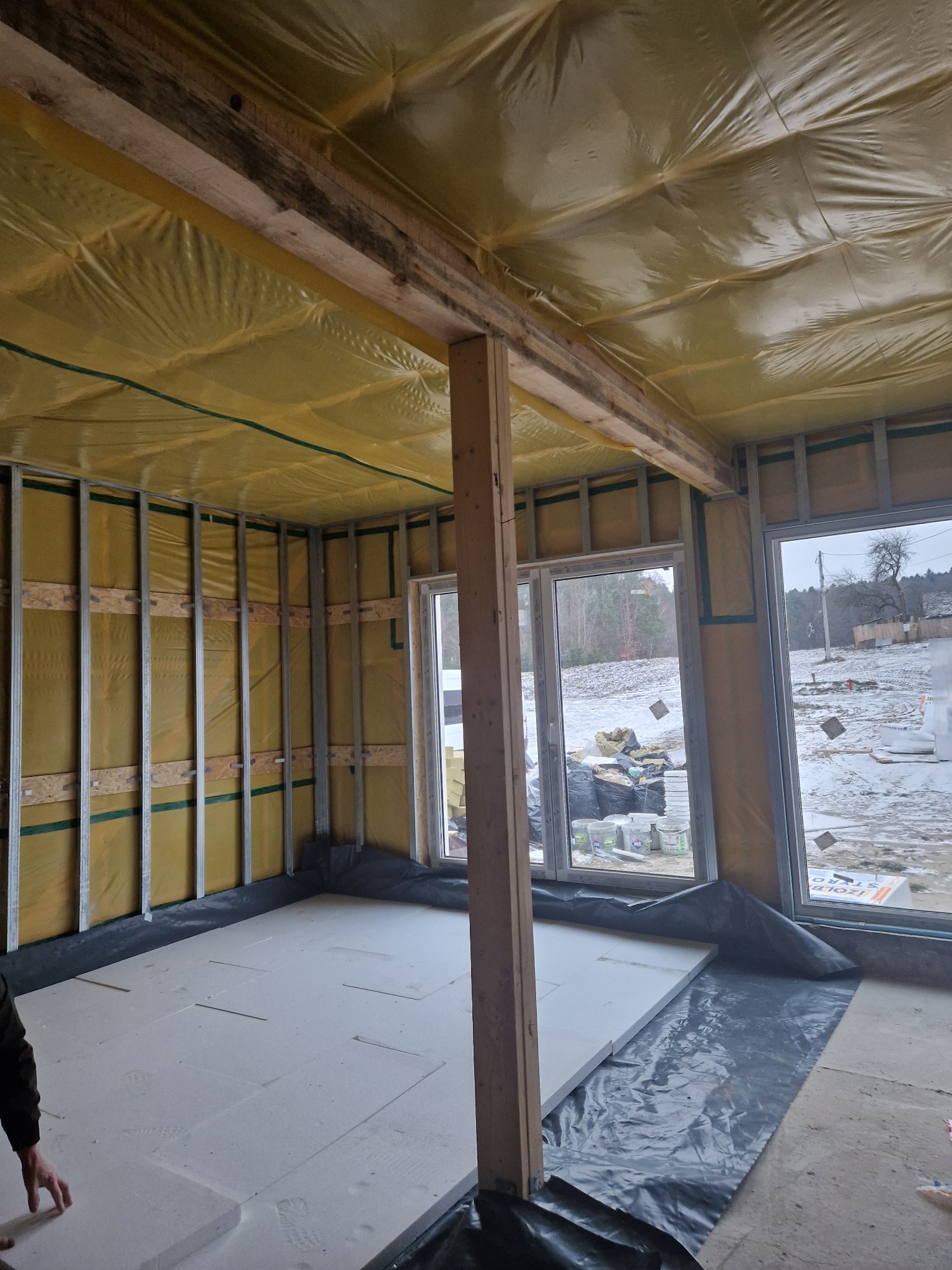 Wnętrze w trakcie budowy: metalowy szkielet ścian, izolacja folią, drewniane belki stropowe, białe płyty izolacyjne na podłodze, okna z widokiem na zimowy krajobraz.