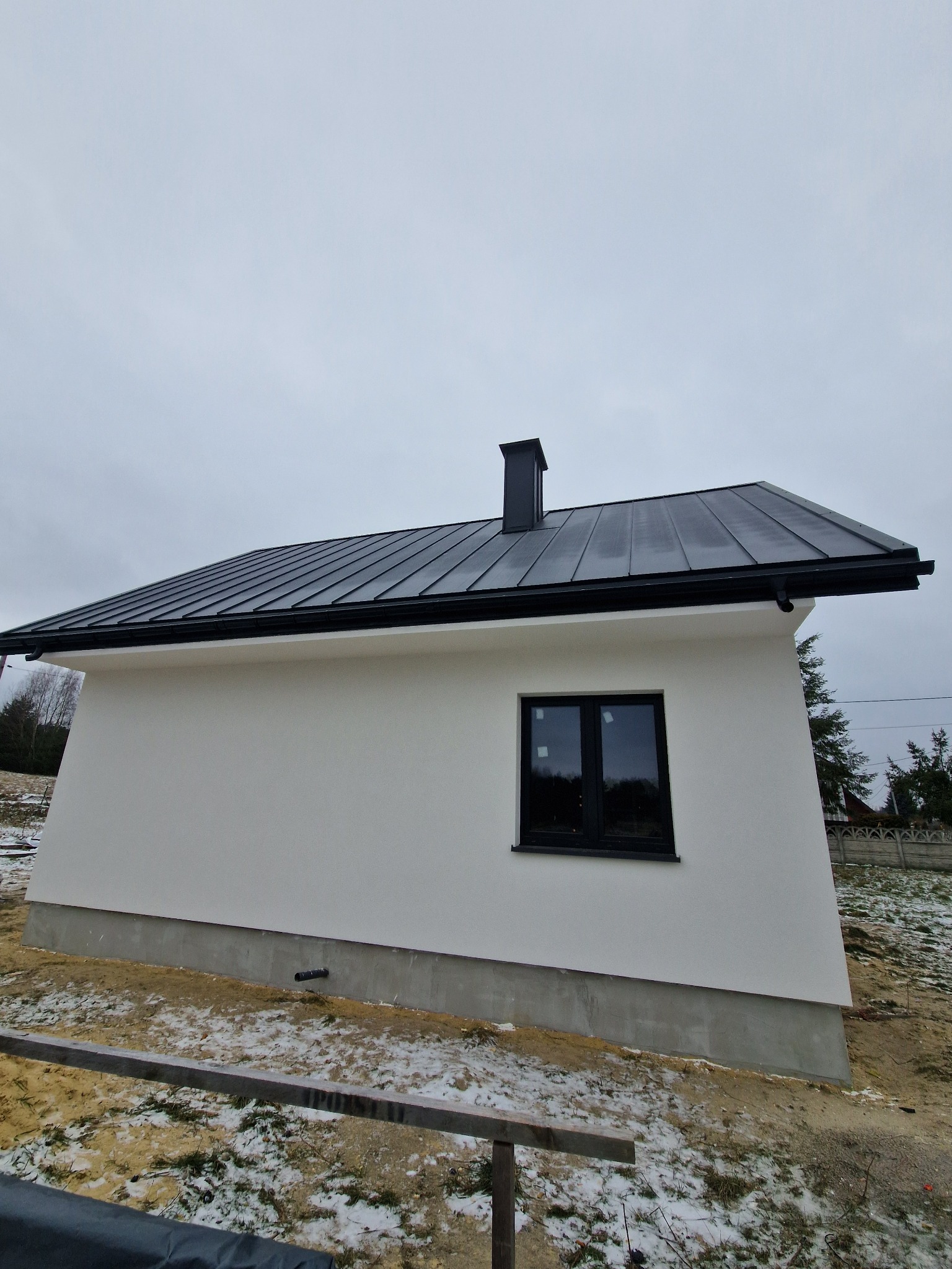 Nowoczesny, parterowy dom z czarnym dachem z blachy na tle pochmurnego nieba. Widoczne okno z czarną ramą i komin. Teren wokół domu częściowo pokryty śniegiem.