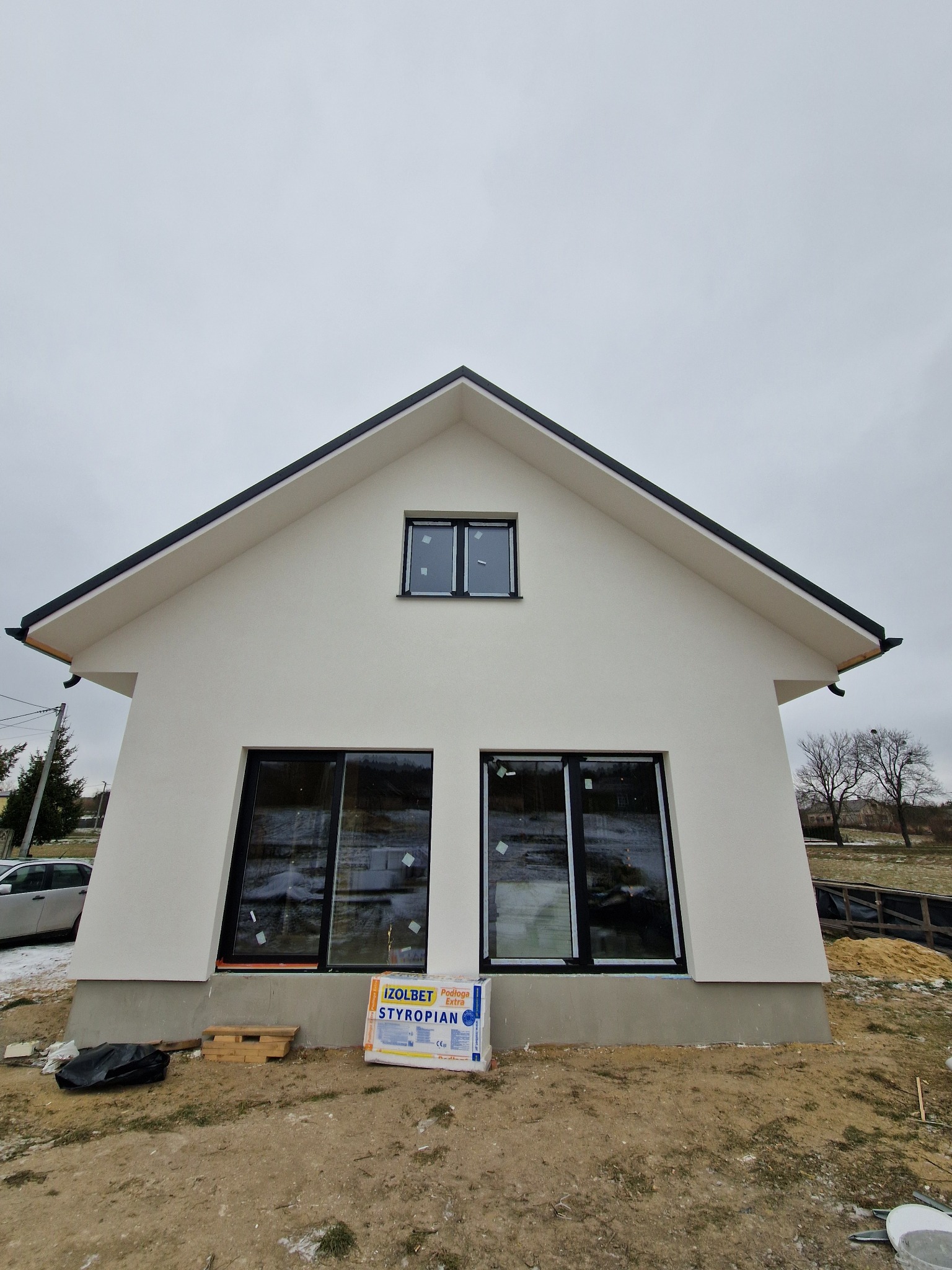 Biały dom z czarnym dachem w trakcie budowy, widoczne okna z folią ochronną i styropian. Ujęcie z dołu, pochmurne niebo w tle.