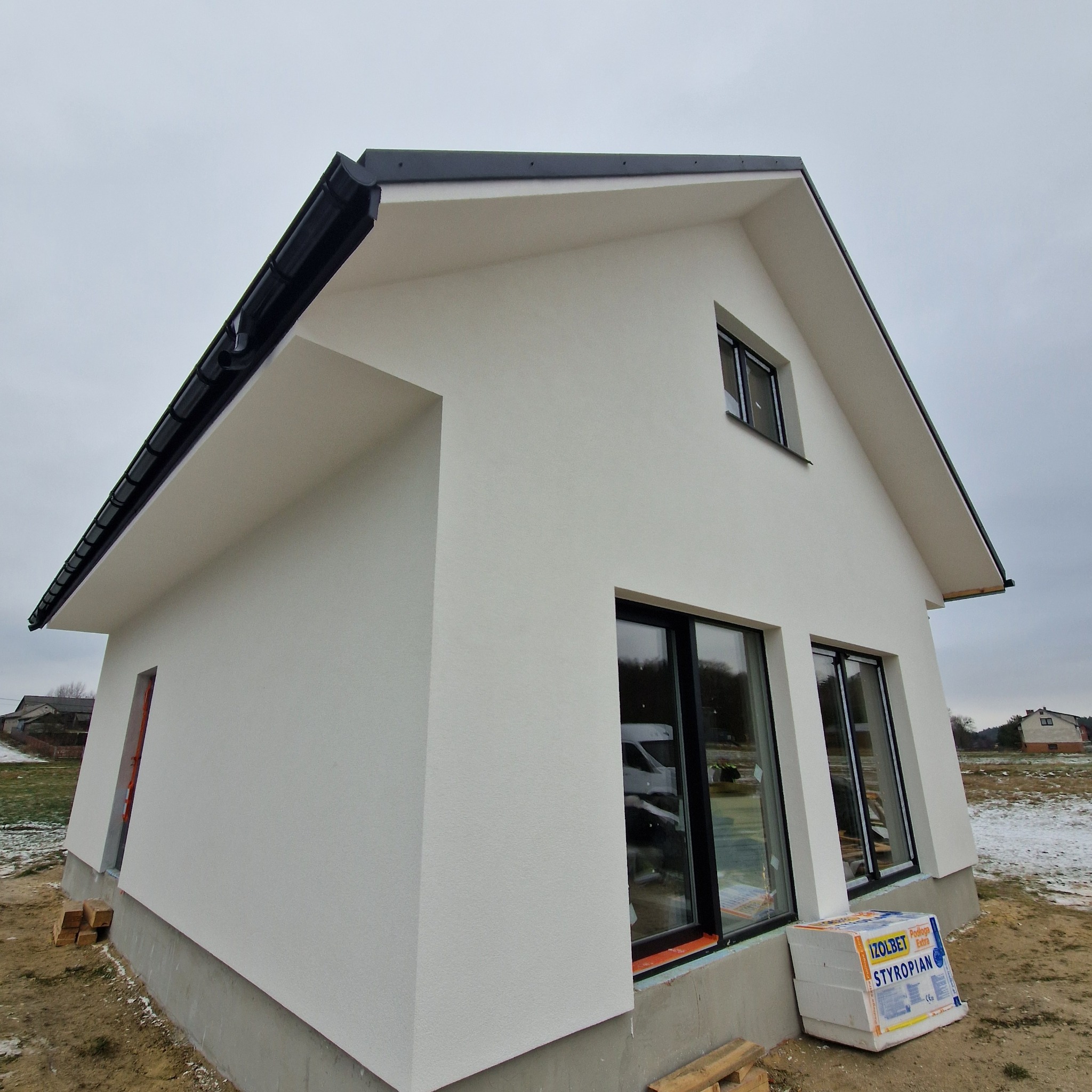 Nowoczesny, biały dom z czarną stolarką okienną i dachem dwuspadowym. Na pierwszym planie paczka styropianu. Widok z boku, pochmurne niebo.