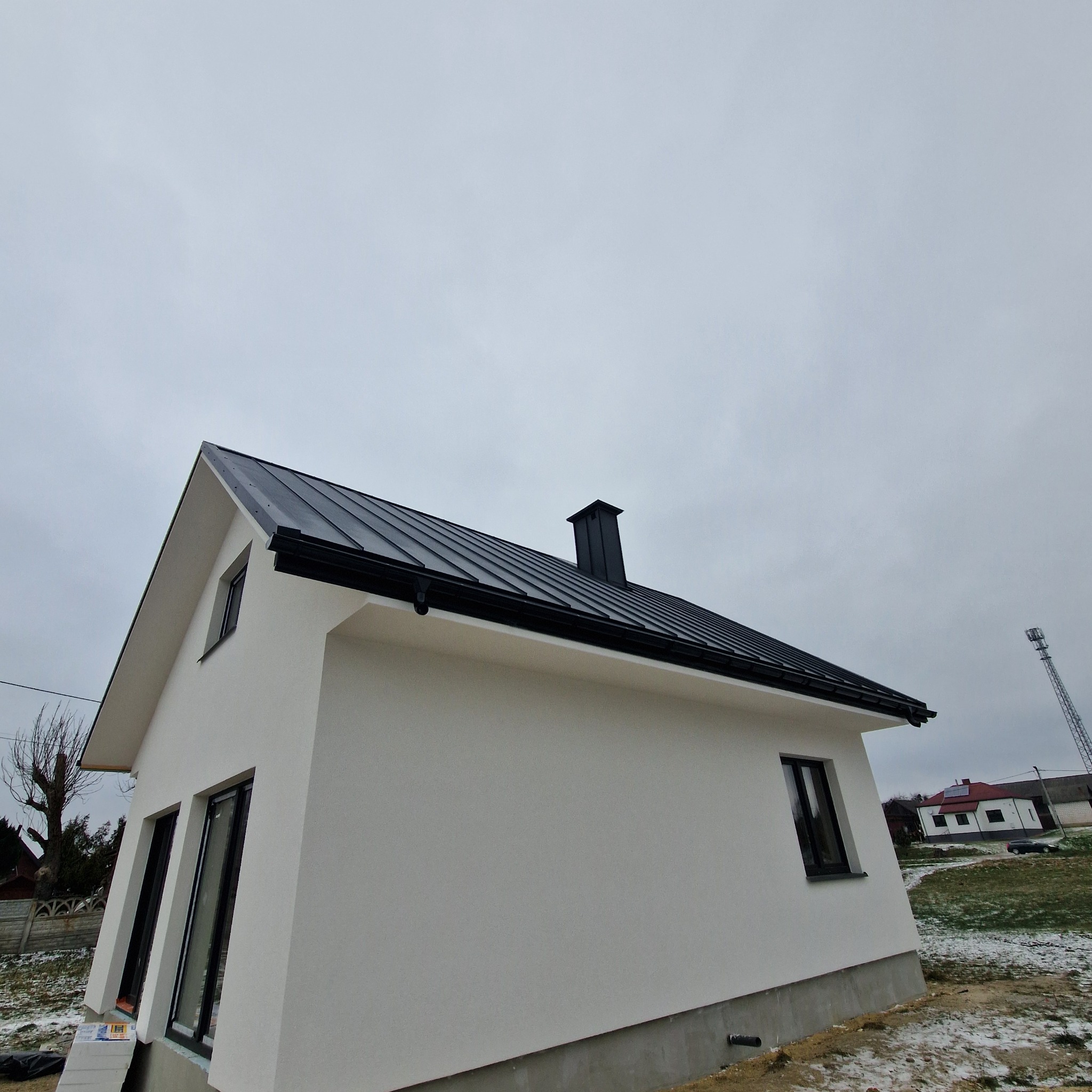 Nowoczesny, biały dom z czarnym dachem z blachy na tle pochmurnego nieba. Widoczny komin i okna. Minimalistyczny styl, świeżo wykończony.