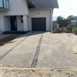 Ułożona kostka brukowa w kolorze szarym, z ciemniejszym pasem wzdłuż podjazdu do garażu nowoczesnego domu jednorodzinnego. Widoczne narzędzia i materiały budowlane.
