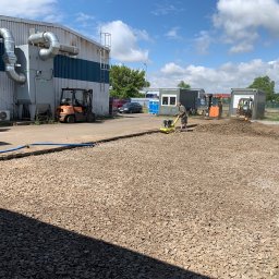 M-bruk Marcin Rybarczyk - Prace przygotowawcze podłoża: zagęszczanie kruszywa zagęszczarką wibracyjną. W tle budynek przemysłowy, kontenery biurowe i sprzęt budowlany.