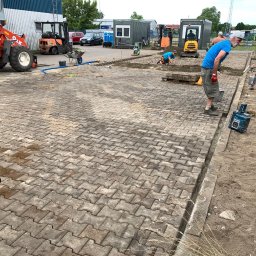 M-bruk Marcin Rybarczyk - Prace brukarskie w toku: robotnicy układają kostkę, widoczne maszyny budowlane, w tym mini koparki i zagęszczarka. Częściowo ułożona nawierzchnia z kostki.