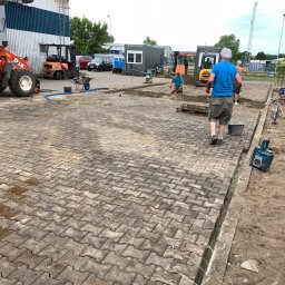 M-bruk Marcin Rybarczyk - Prace brukarskie w toku: robotnicy układają kostkę na placu z użyciem mini-koparki i zagęszczarki. Część kostki już ułożona, widoczne narzędzia i materiały budowlane.