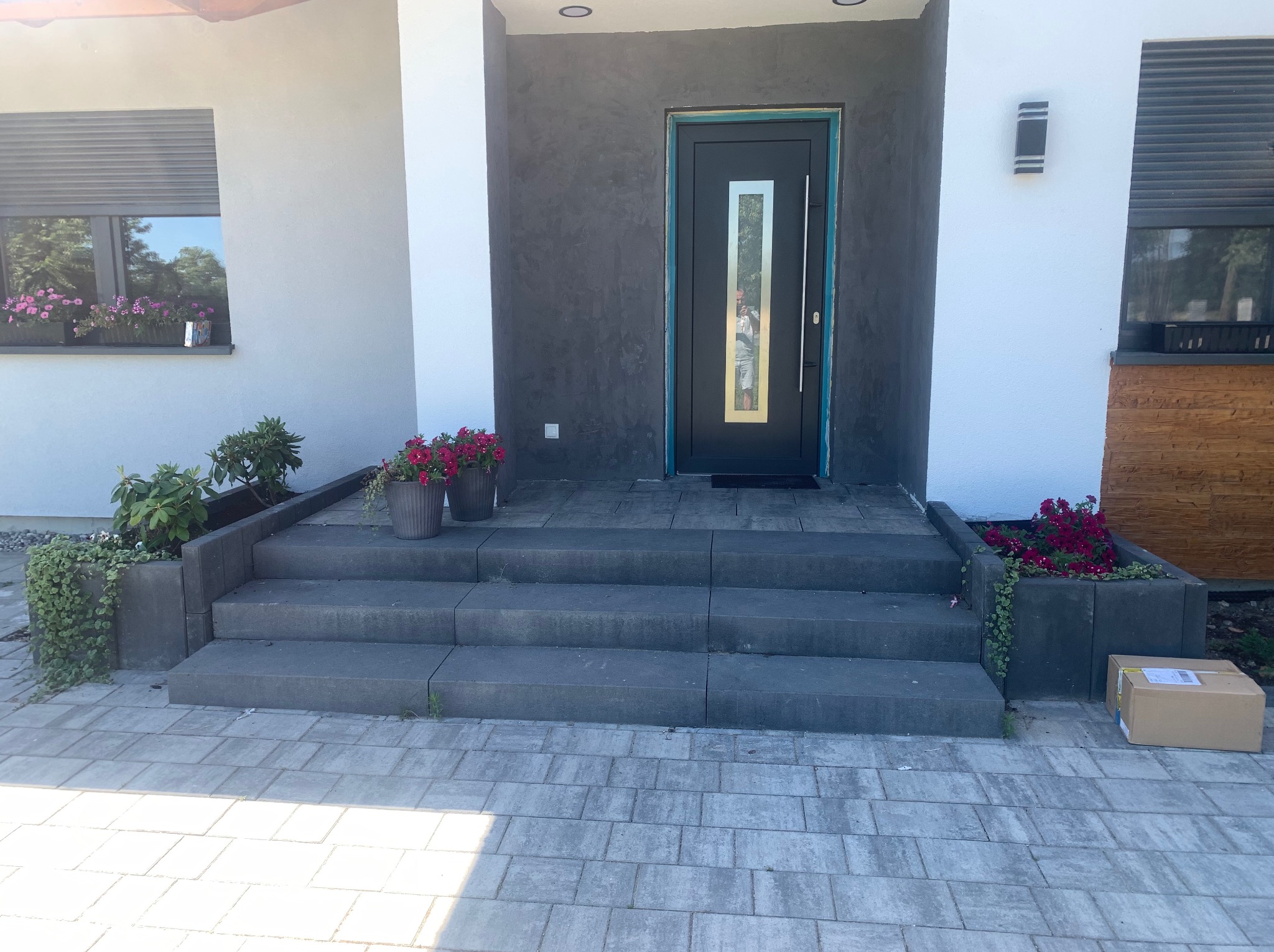 Nowoczesne wejście do domu z kostki brukowej, schody z donicami z kwiatami, czarne drzwi wejściowe z przeszkleniem. Paczka leży na bruku.