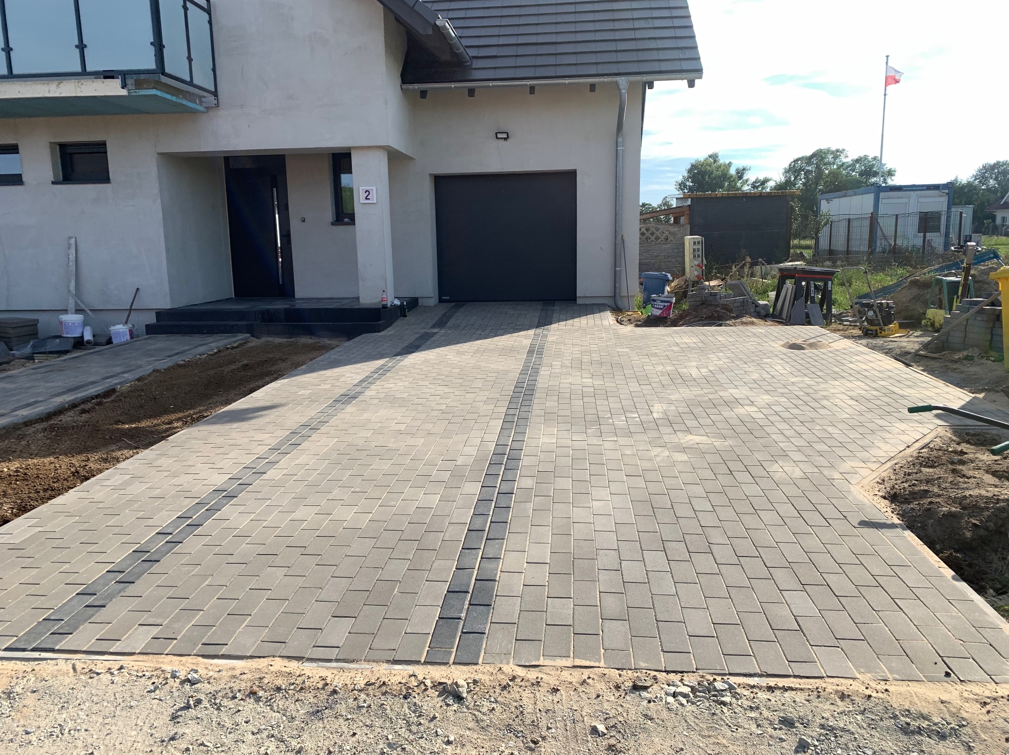 Ułożona kostka brukowa w kolorze szarym, z ciemniejszym pasem wzdłuż podjazdu do garażu nowoczesnego domu jednorodzinnego. Widoczne narzędzia i materiały budowlane.