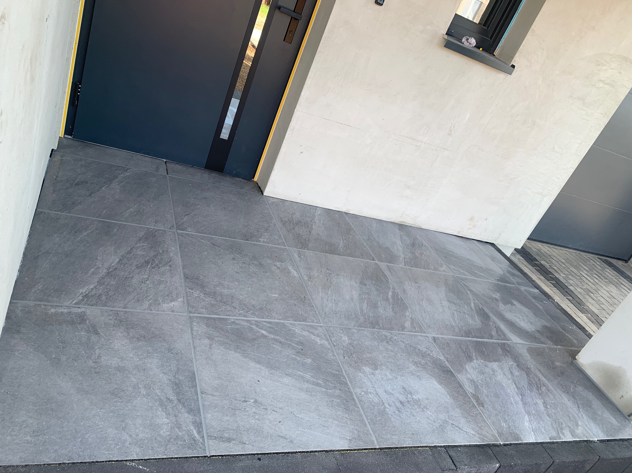 Ułożona, grafitowa płytka gresowa na zewnątrz budynku, widoczna przy wejściu i garażu. Płytki o fakturze kamienia, ułożone równo, z widocznymi fugami.