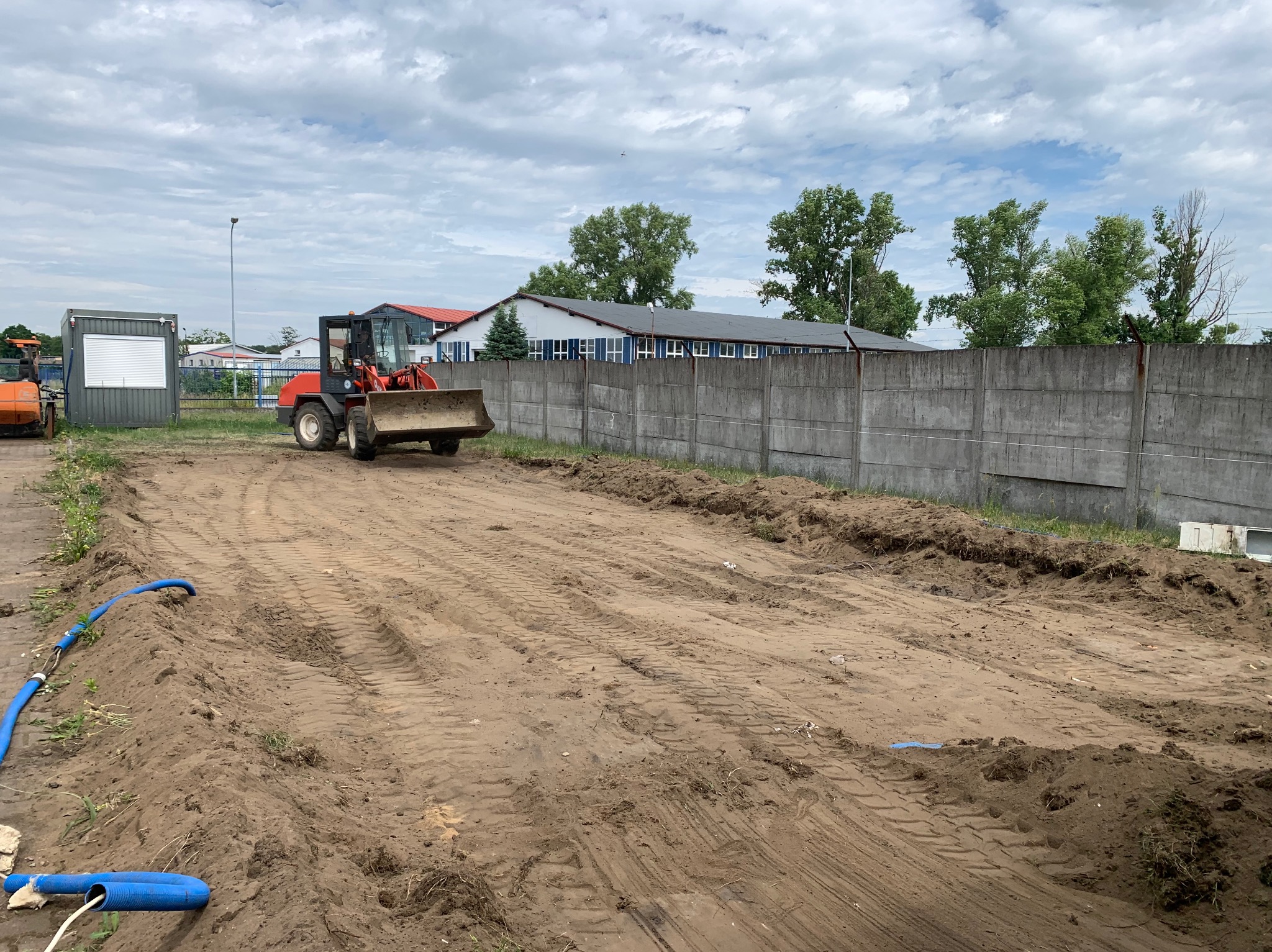 Niwelacja terenu w Deszcznie: Czerwona ładowarka kołowa wyrównuje ziemię na placu budowy. Widoczne ślady opon i betonowe ogrodzenie.