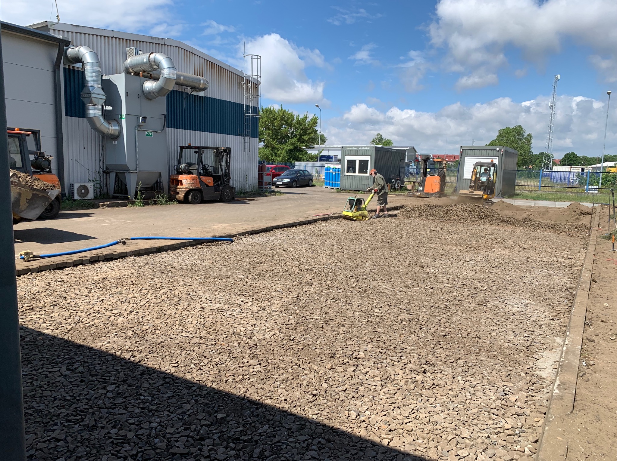 Prace przygotowawcze podłoża: zagęszczanie kruszywa zagęszczarką wibracyjną. W tle budynek przemysłowy, kontenery biurowe i sprzęt budowlany.