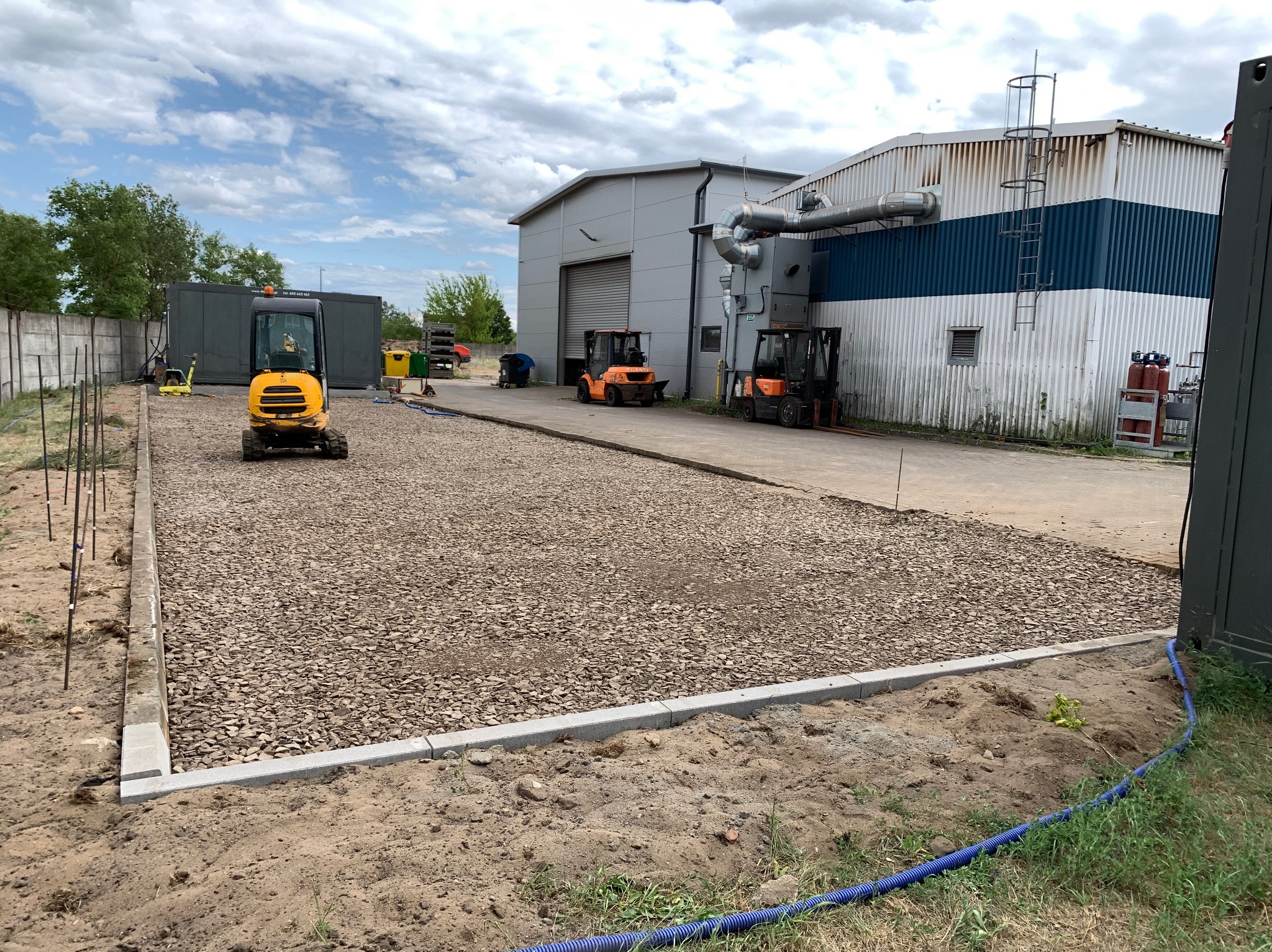 Przygotowany teren pod kostkę brukową z obrzeżami, widoczny mini-koparka i maszyny budowlane. Częściowo wysypany gruz. Widok na budynek przemysłowy.